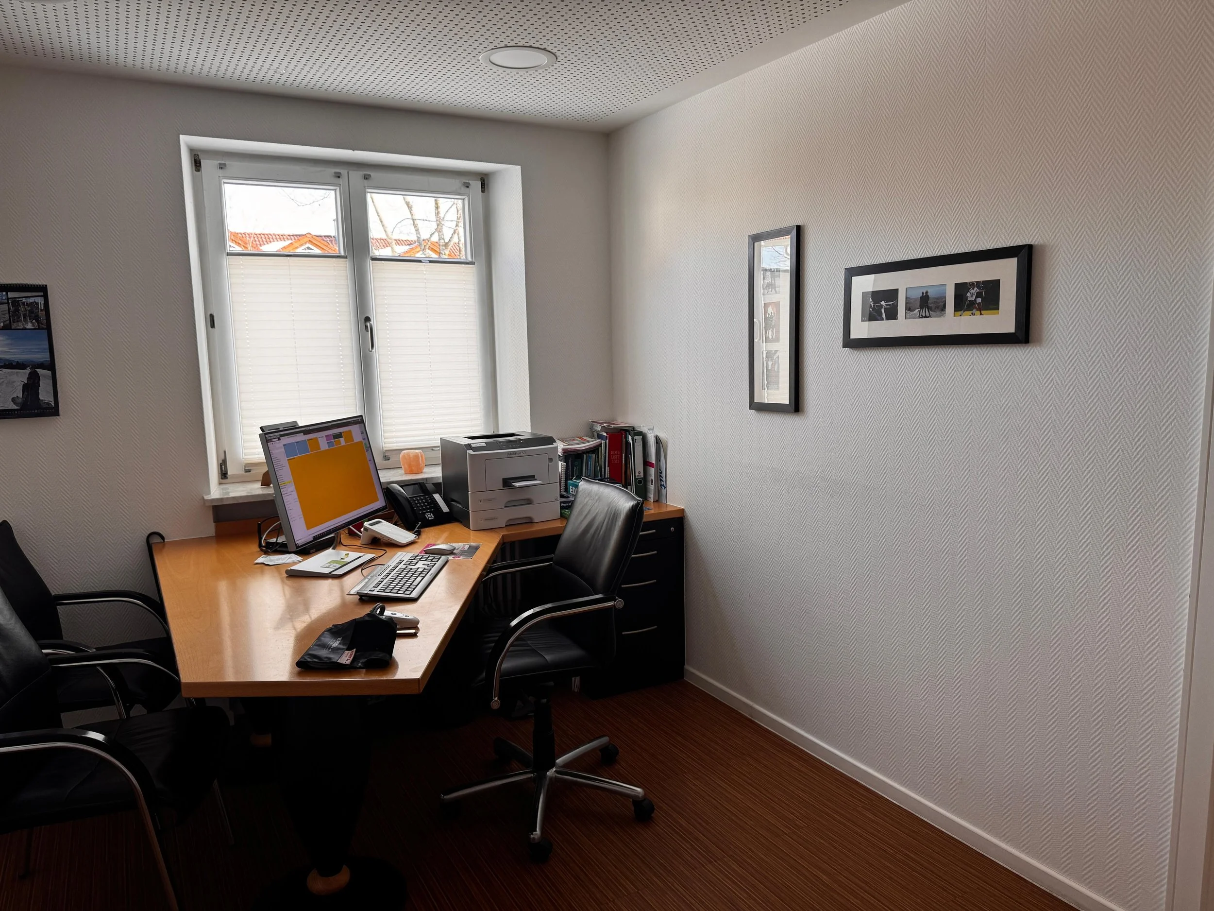 Ein kleines Büro mit einem Holz-Schreibtisch, auf dem ein Computerbildschirm, eine Tastatur, ein Telefon, ein Drucker, einige Aktenordner und Schreibwaren stehen. Es gibt drei schwarze Stühle, eine Arbeitsplatzlampe und eine Fenster mit weißen Fensterläden, durch die ein Blick auf Dächer und Bäume außerhalb möglich ist.