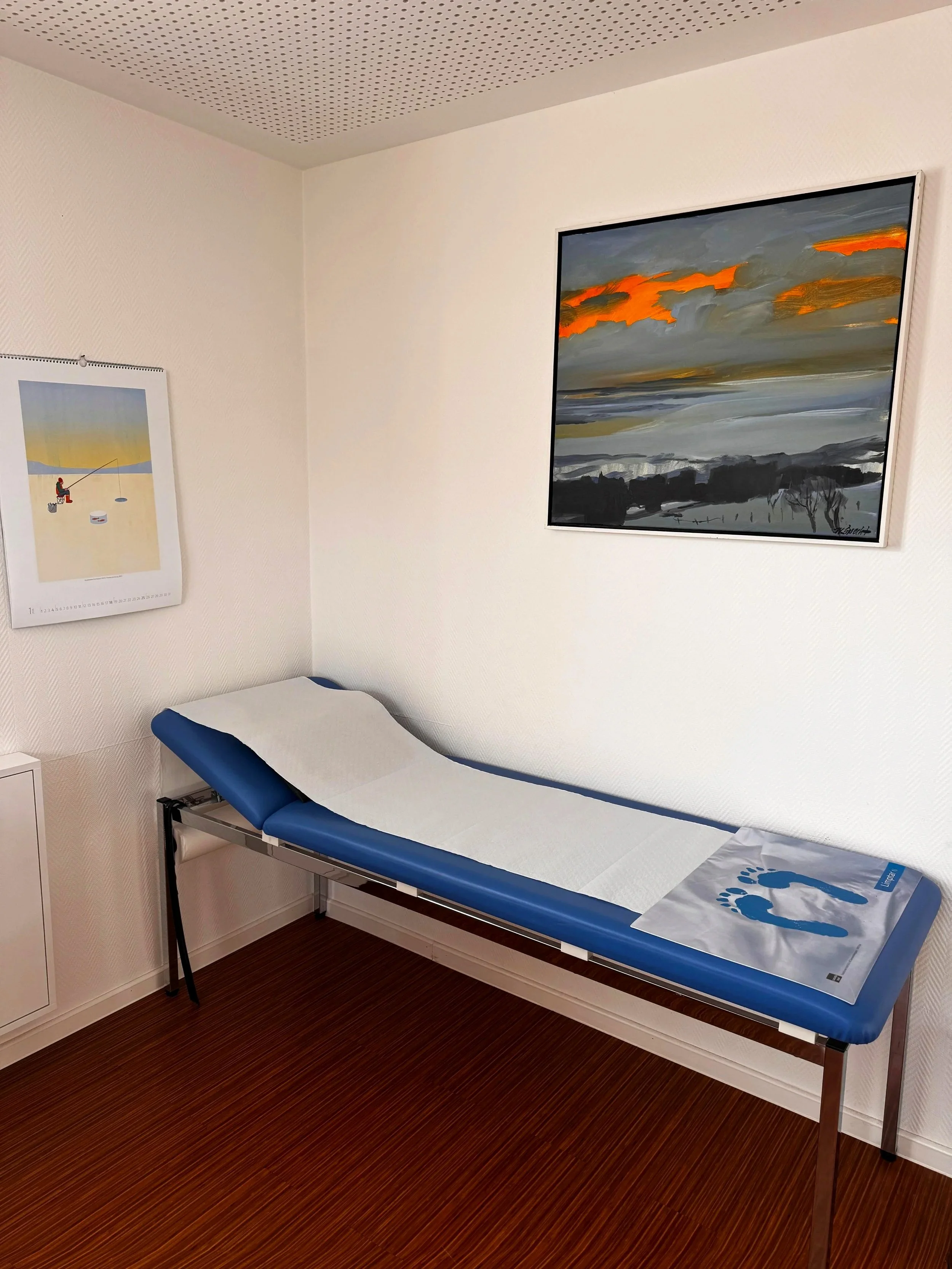 ein Untersuchungstisch in einer medizinischen Praxis mit blauer Polsterung, weißen Papierdeckel und einer Informationskarte mit Fußabdrücken