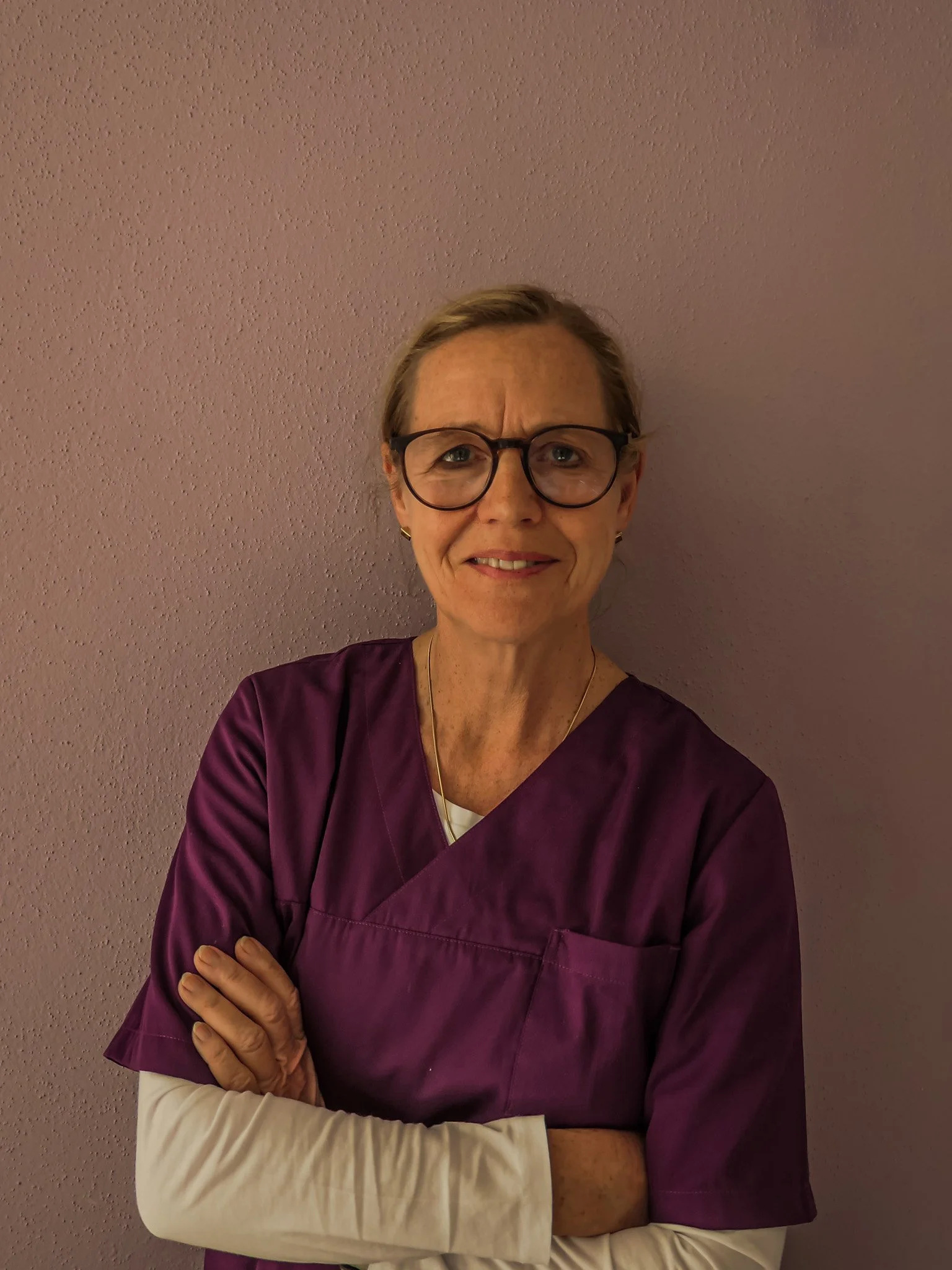 Eine ältere Frau trägt eine violette medizinische Arbeitskleidung und schwarze Brille. Sie steht vor einer helllila Wand mit verschränkten Armen.