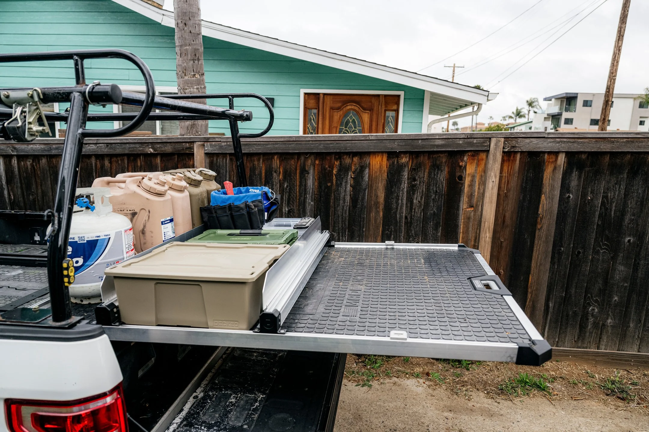 Back of a utility truck equipped with DECKED CargoGlide with gardening supplies including gas cans, propane tank, and storage containers, parked in a yard enclosed by a wooden fence with a green house and palm trees in the background.
