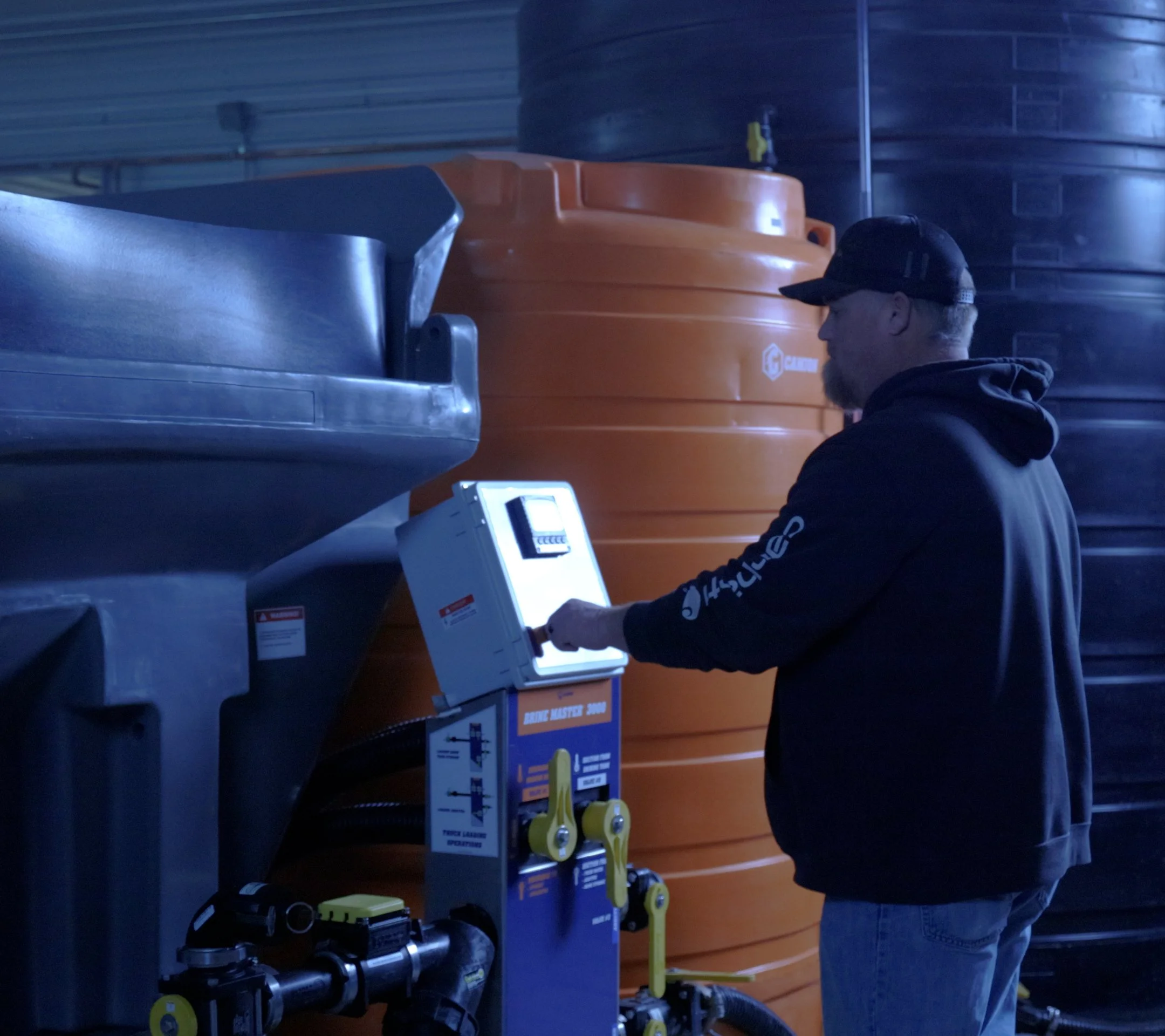 Man with a beard and cap operating a brine machine control panel in a dark industrial setting with storage tanks in the background.