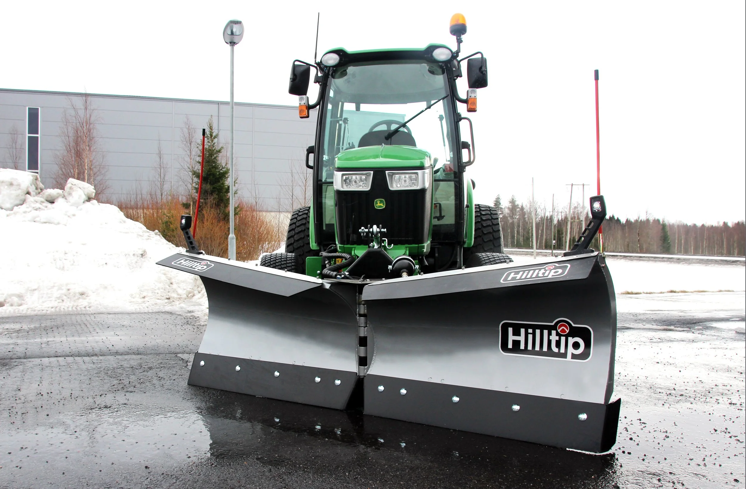 Green tractor equipped with a large snow plow blade attached to the front, clearing snow from a paved surface outdoors.