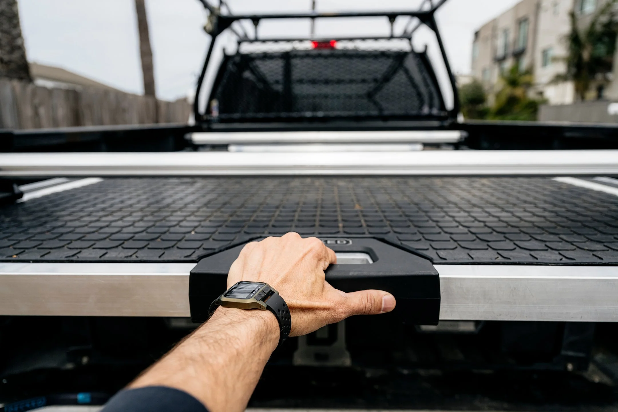 A person's hand holding a handle on a CargoGlide installed in a pickup truck, with the truck's bed and a black metal rack visible, in an outdoor area with houses and trees in the background.