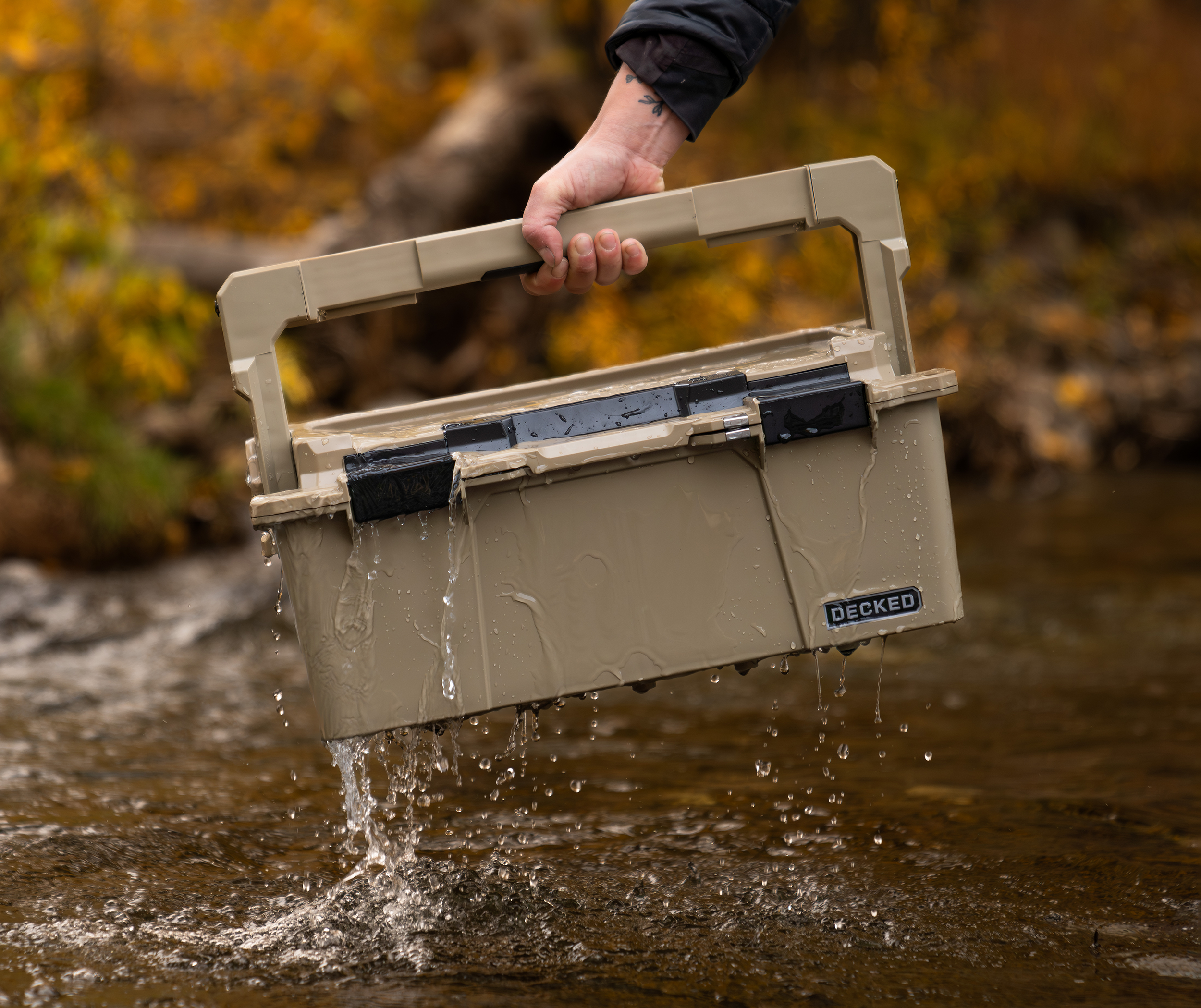 Person holding a beige waterproof D-Co storage container over a shallow stream during autumn.