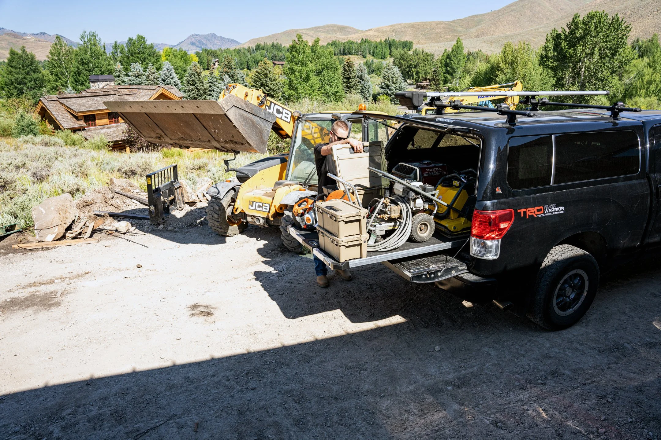 Construction workers preparing equipment from a pickup truck equipped with DECKED CargoGlide near a yellow compact excavator at a construction site in a mountainous area.