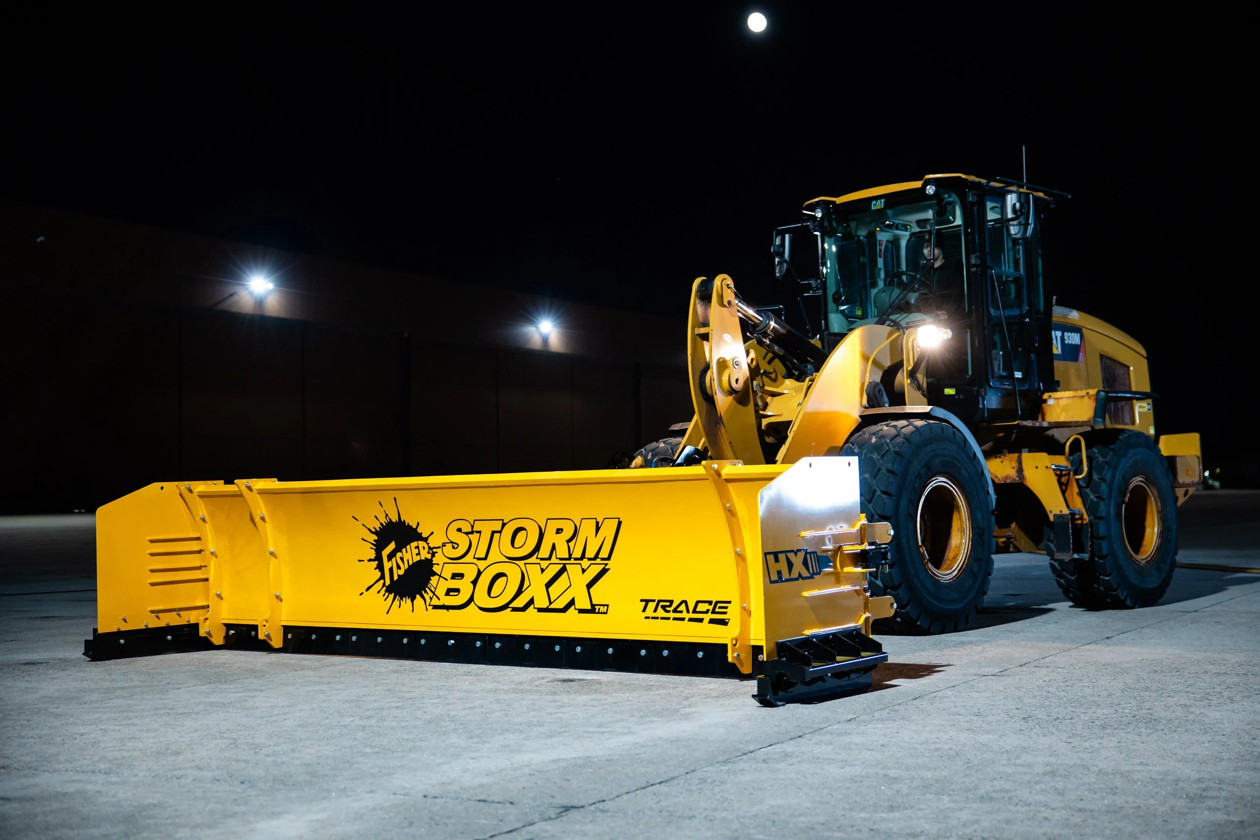 A yellow Fisher Storm BoXXX snow blower attached to a yellow tractor, operating at night outside under bright lights.