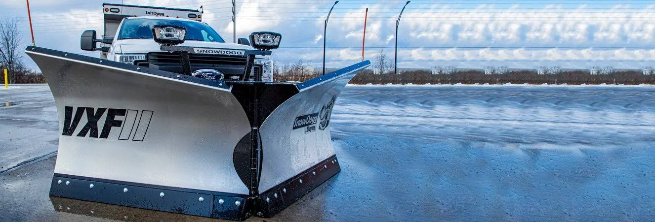 A white pickup truck equipped with a snow plow attachment on the front, parked on a wet frozen surface with a cloudy sky in the background.