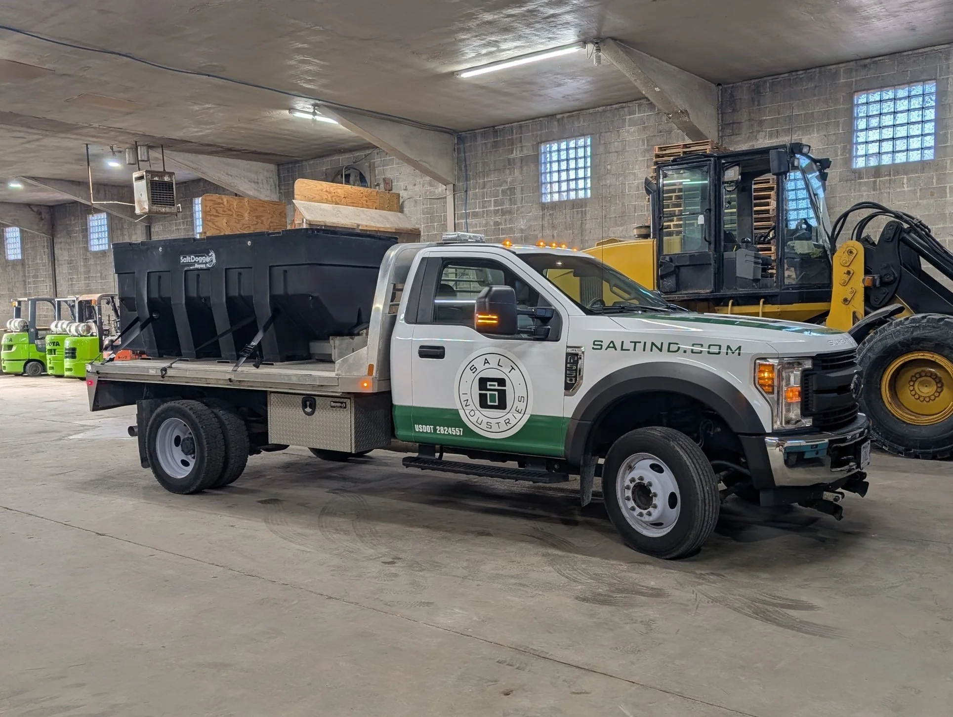 A white and green Salt Industries service truck with a black salt spreader loaded in the back parked inside a warehouse, with heavy machinery and green equipment in the background.