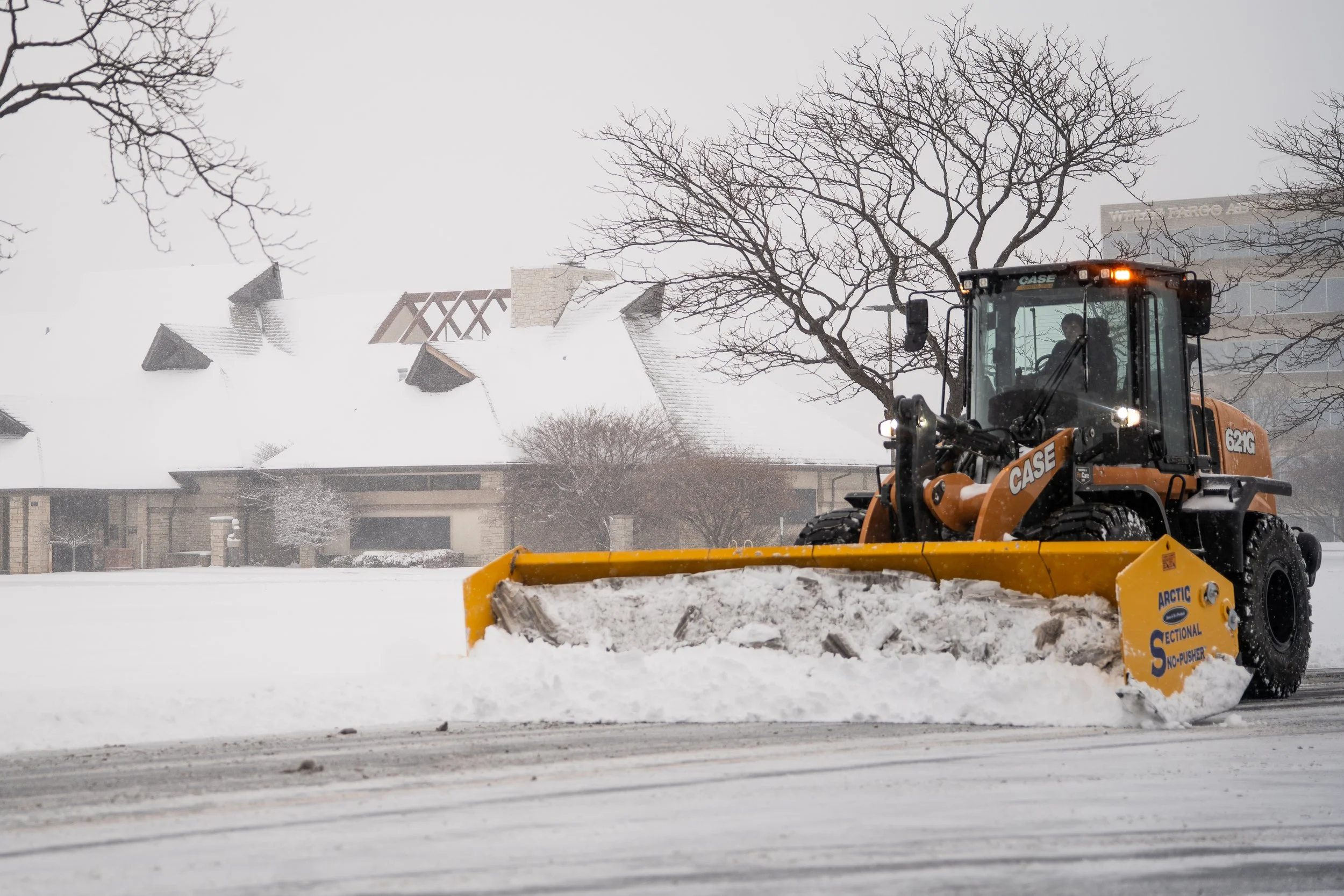A Fisher StormBoxx clearing snow from a road with houses, trees, and a shopping center in the background on a snowy day.