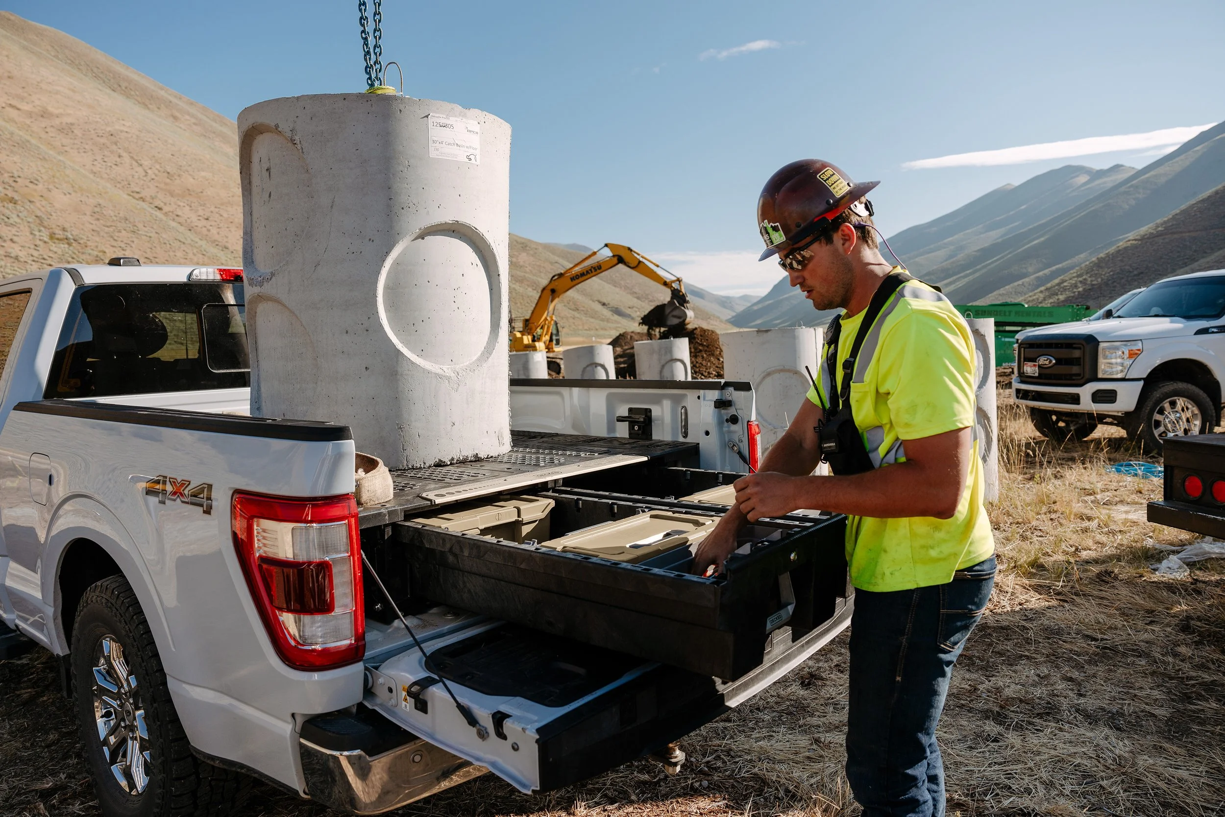 Construction worker in a yellow safety vest and helmet working at a construction site with concrete cylinders on the back of a pickup truck equipped with DECKED Drawers, mountains in the background.