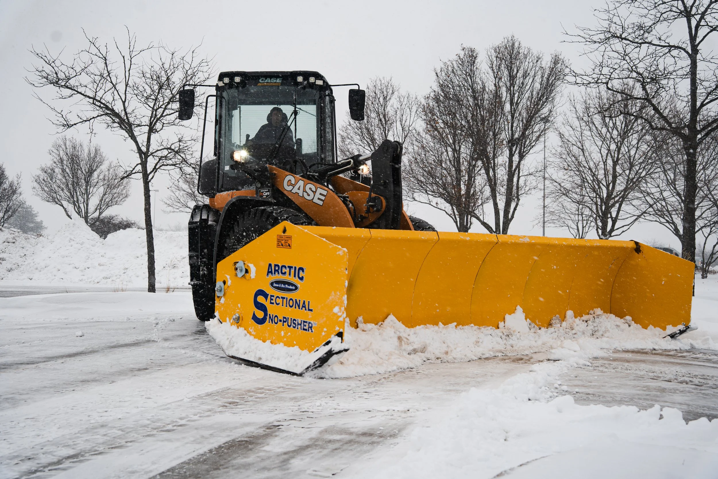 A person operating a sectional ARCTIC push box on a snowy road during winter, with leafless trees in the background.