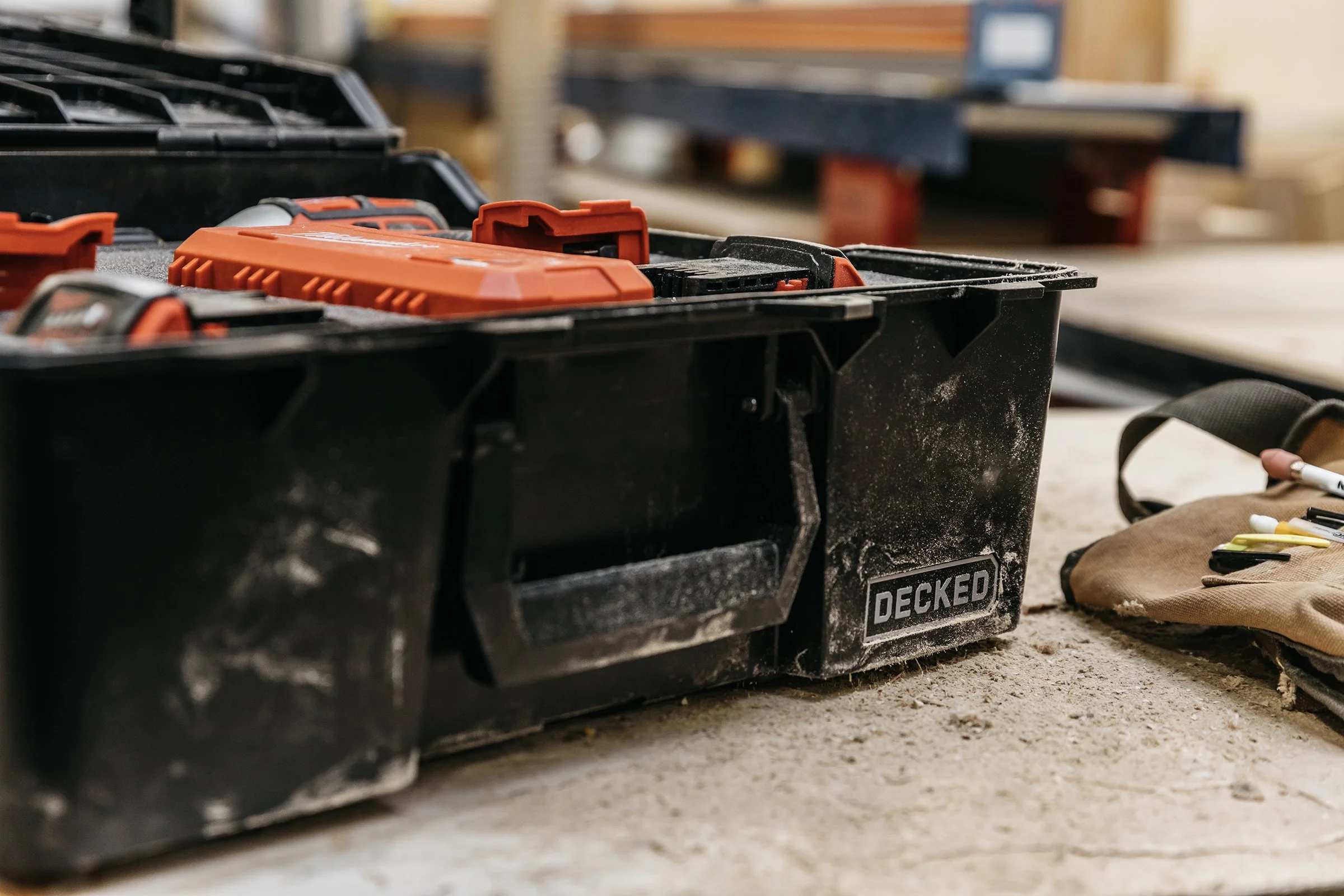 A black D-Co case with a red power drill inside, sitting on a dusty workbench in a woodworking shop.