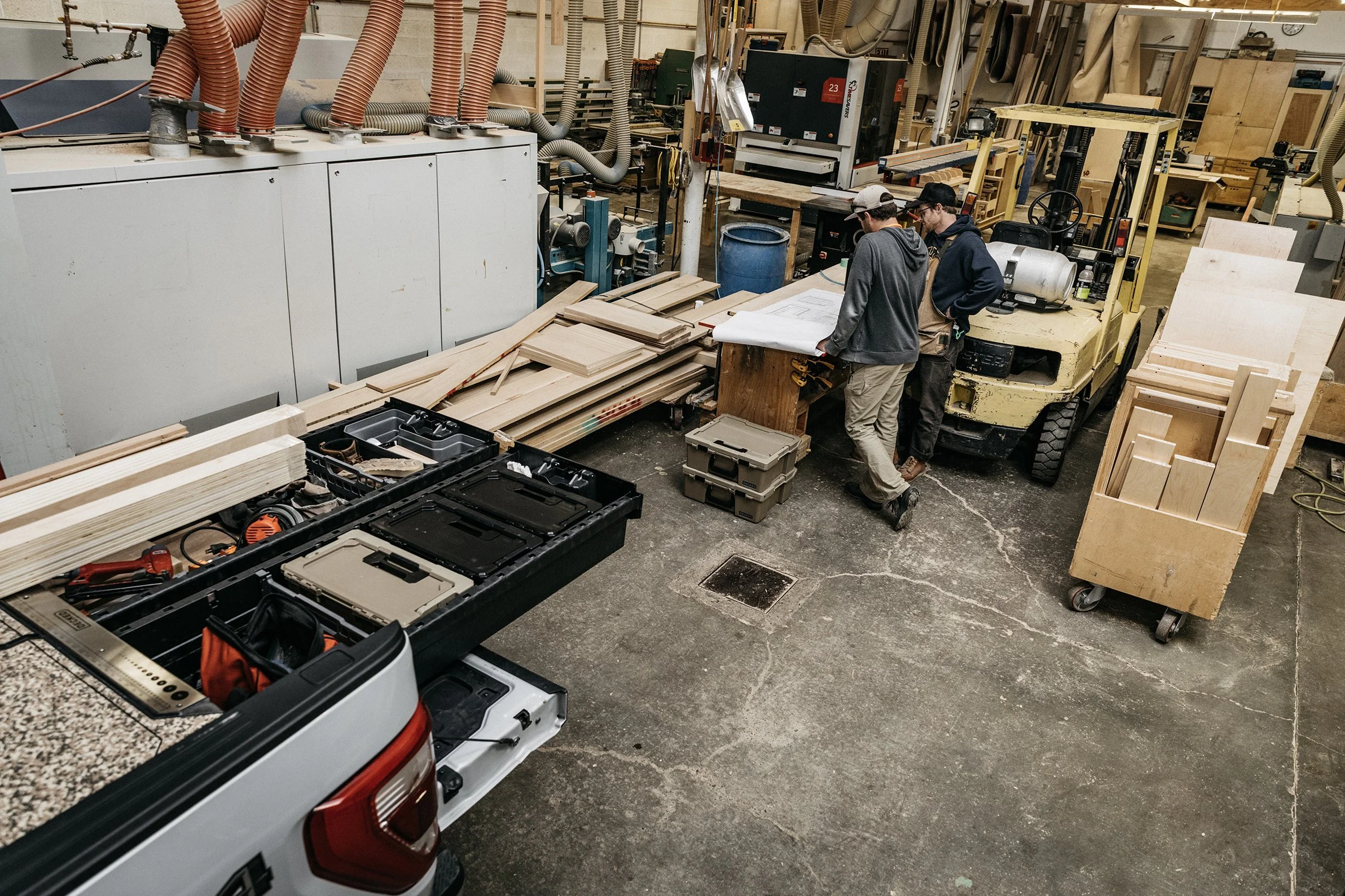 Three people working in a woodworking shop, standing near a workbench with a pick-up truck equipped with DECKED Drawers and D-Co Cases