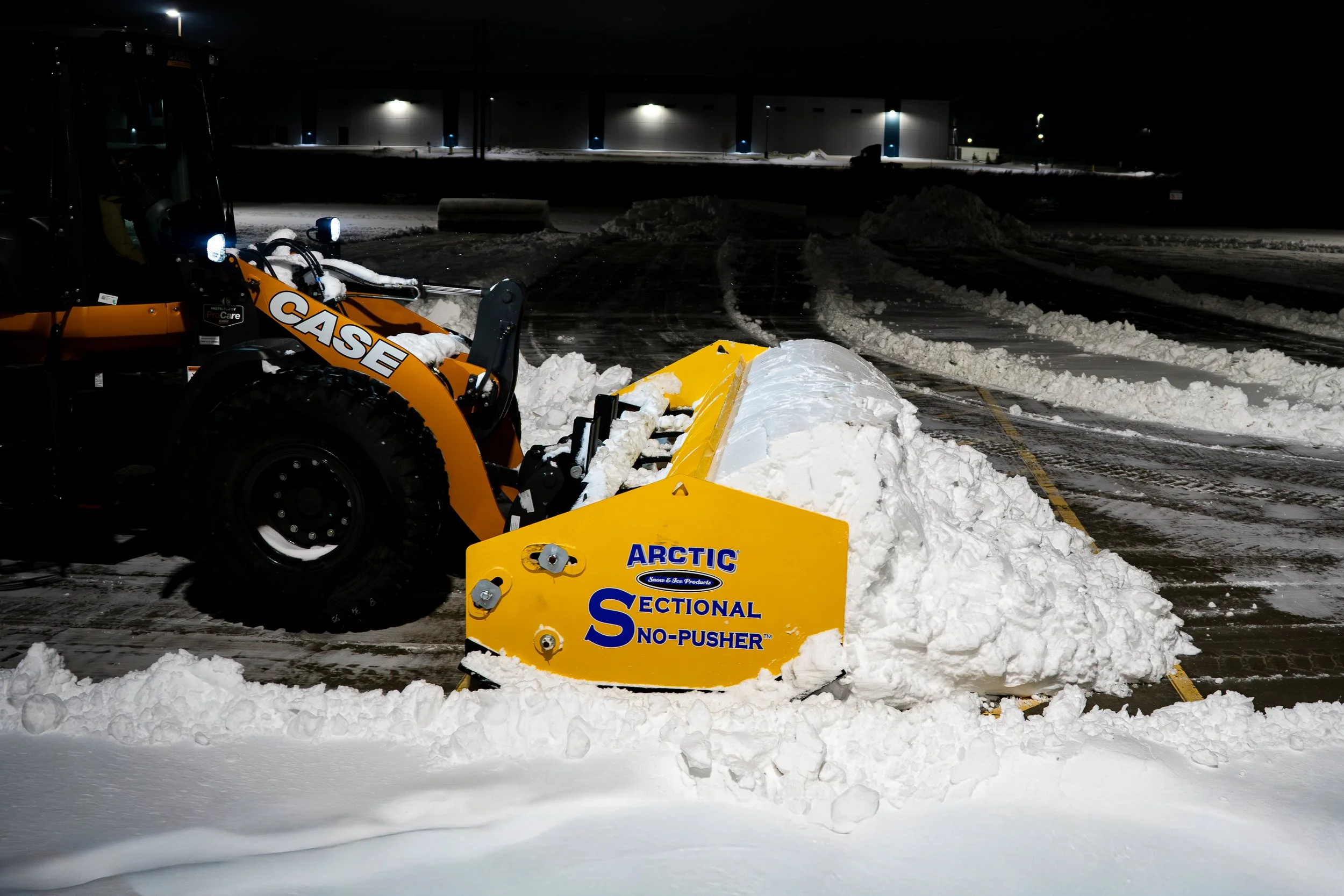 An Arctic snow pusher clearing snow in a parking lot at night, with snow piles and a large building in the background.
