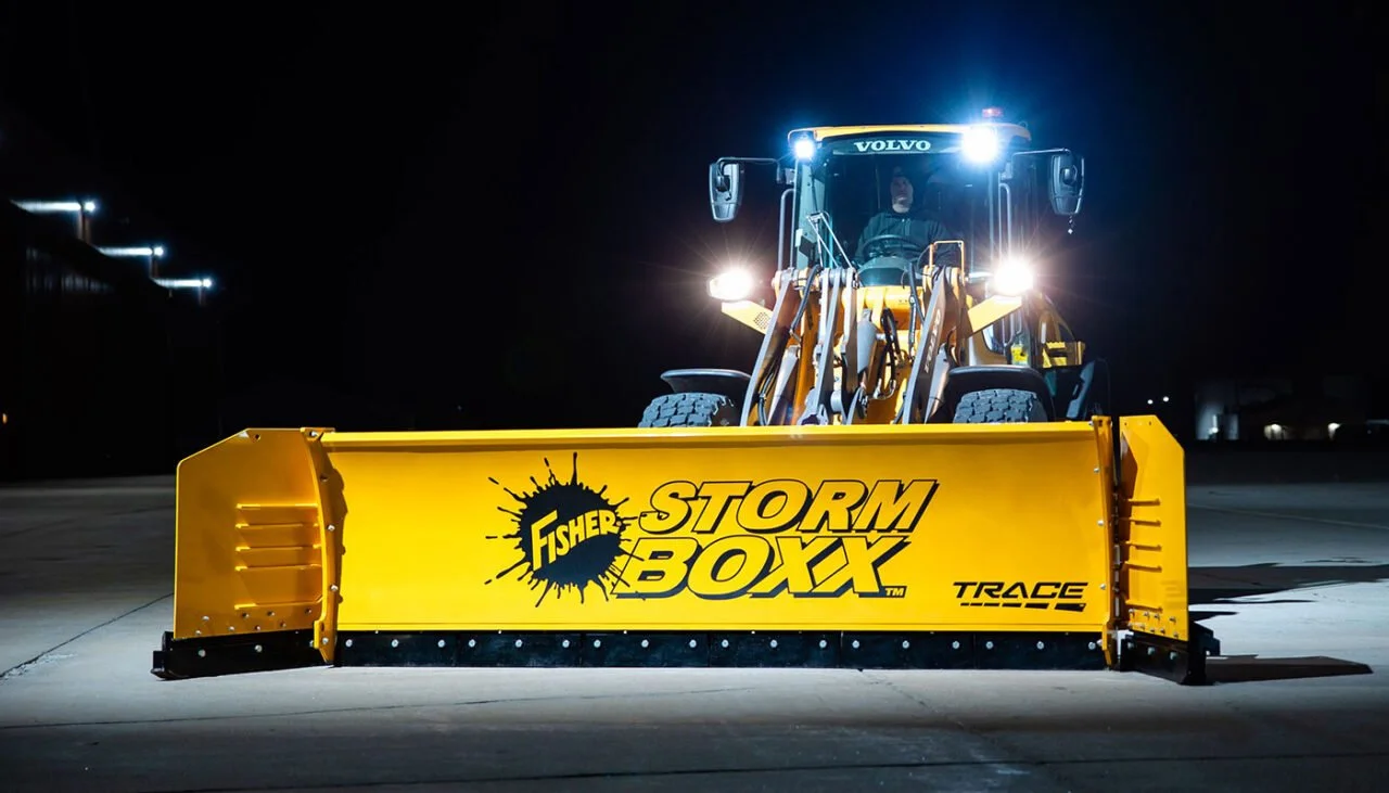 A yellow snow plow with the logo Fisher Storm Boxx on the front, attached to a Volvo construction vehicle, is positioned on pavement at night, with the vehicle's lights on.