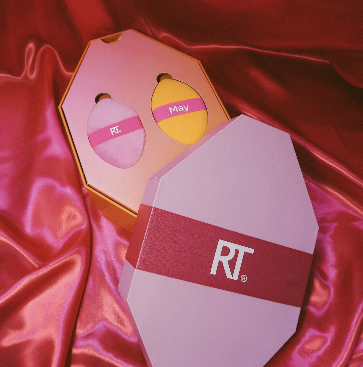 A pink and yellow box of branded skincare products, with two round containers labeled "RT" and "May" inside a pink hat-shaped opening, placed on shiny pink satin fabric.