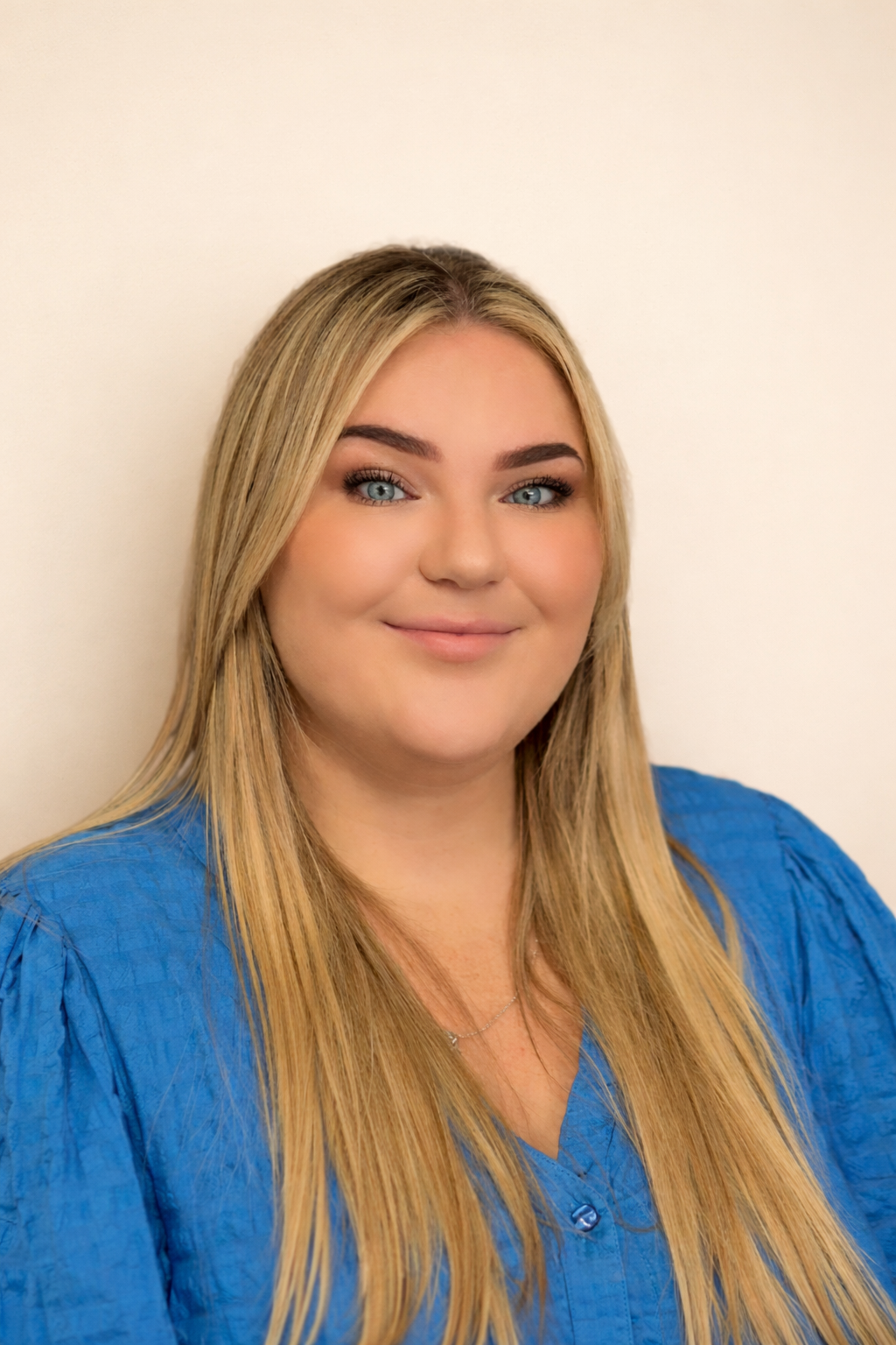 A woman with long blonde hair, blue eyes, and a subtle smile, wearing a blue top, standing against a dark purple brick wall.