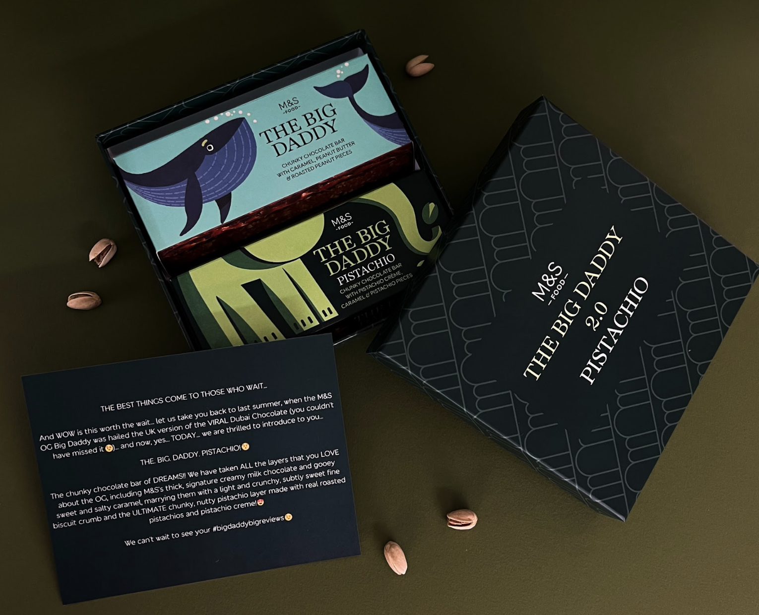 A black gift box with white decorative lines containing two M&S Food 'The Big Daddy' chocolate bars, one with a whale design labeled 'Whale Big Daddy' and the other with a pistachio design labeled 'Pistachio Big Daddy.' The box is open, placed on a dark surface with scattered pistachio nuts and a black note card with white and pink text, describing the chocolate bars.