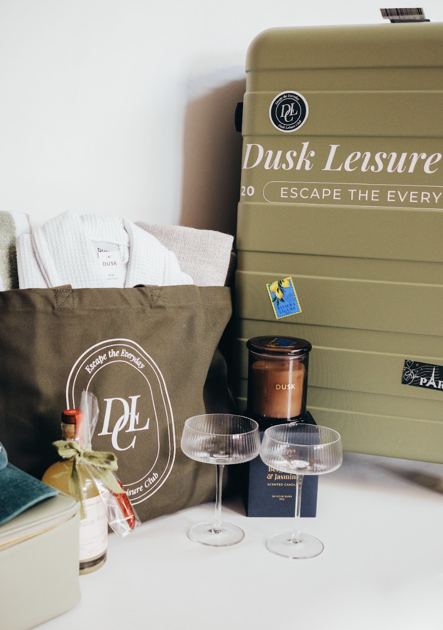 Collection of home and travel items including a large green suitcase, a fabric tote bag with a logo, two coupe glasses, a candle, a small box, a bottle with a ribbon, and some linens in a basket.