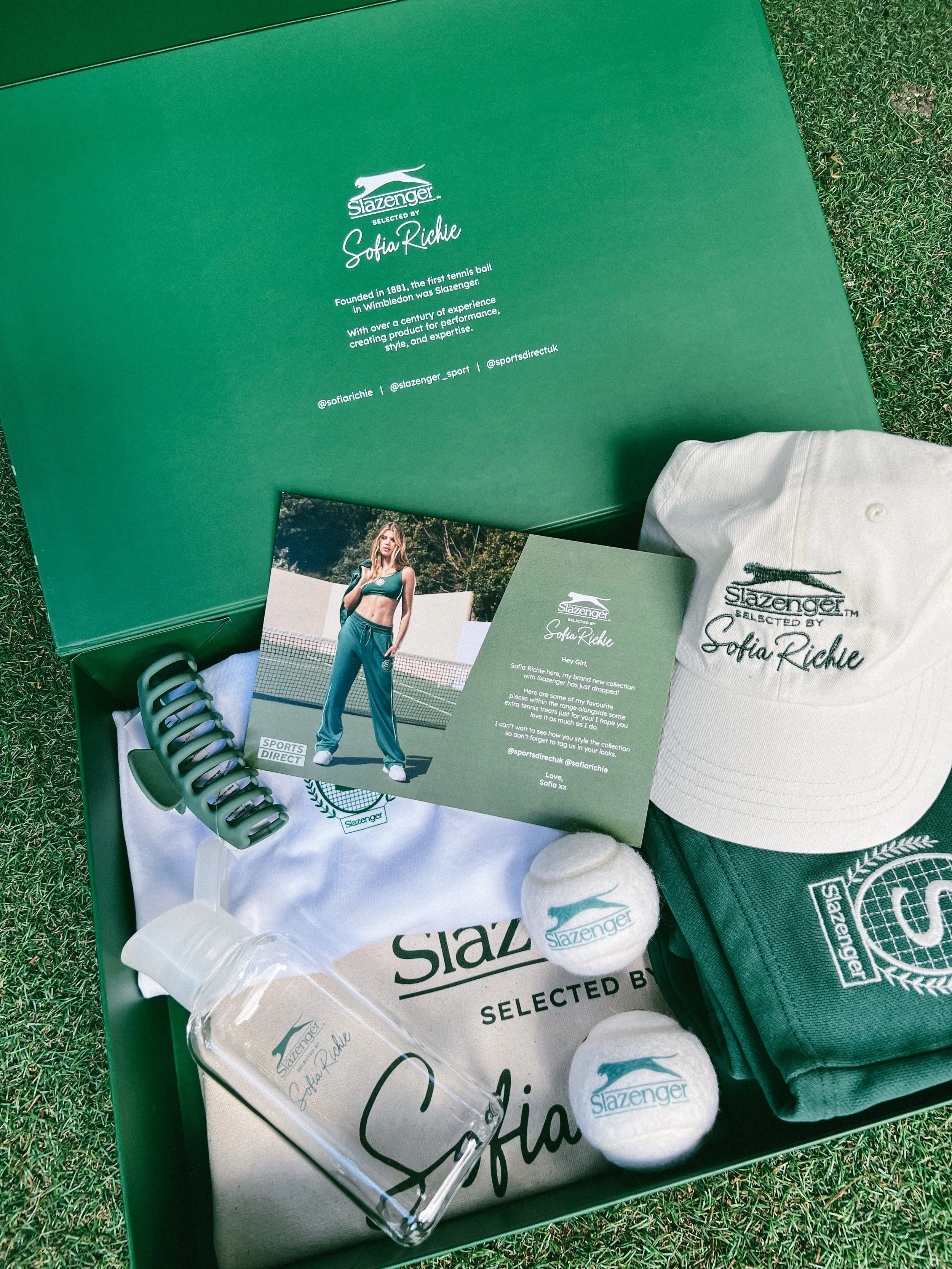 Box containing Slazenger tennis equipment and apparel, including two tennis balls, a water bottle, cap, towel, and a promo card with a tennis player, on grass.