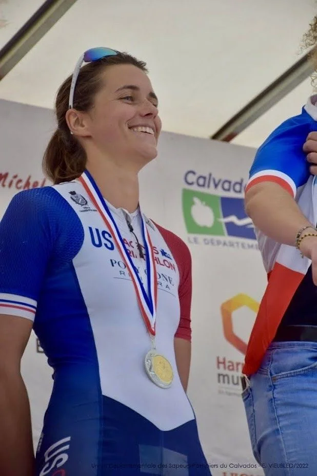 Une femme souriante portant une médaille d'or, vêtue d'une tenue de sport bleue, blanche et rouge, lors d'une cérémonie de remise de prix.