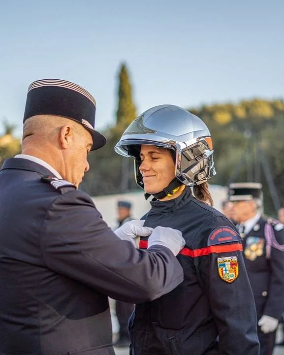 Un officier en uniforme remet une médaille à une femme portant un casque de moto, lors d'une cérémonie officielle en plein air.