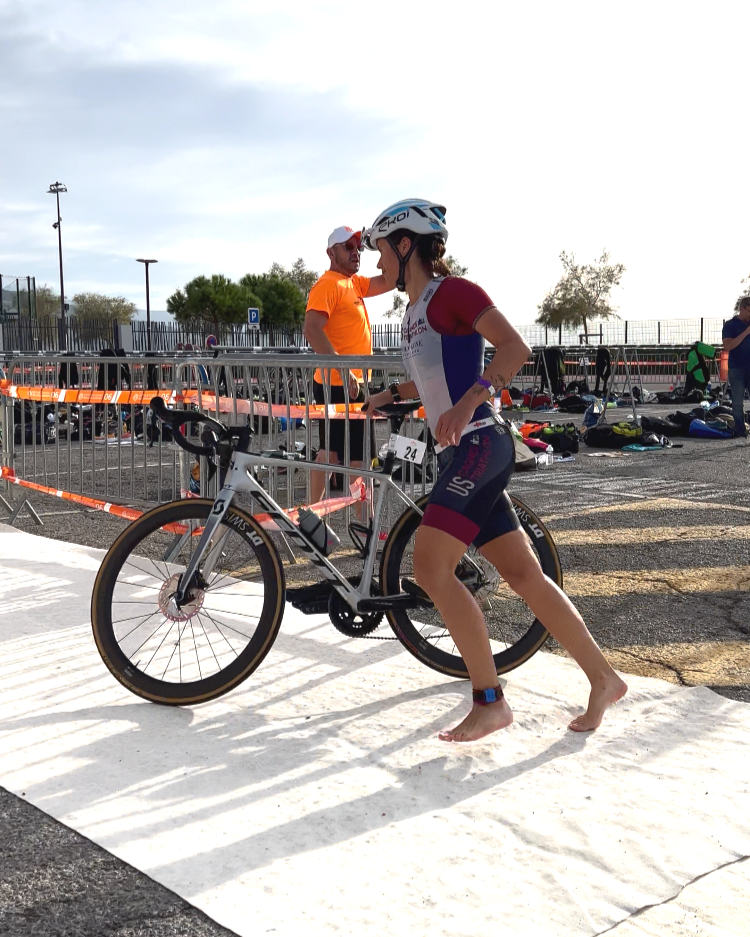 Une femme en équipement de cyclisme pousse son vélo mudérisé lors d'une compétition outdoor, avec d'autres participants et équipements de sport en arrière-plan.