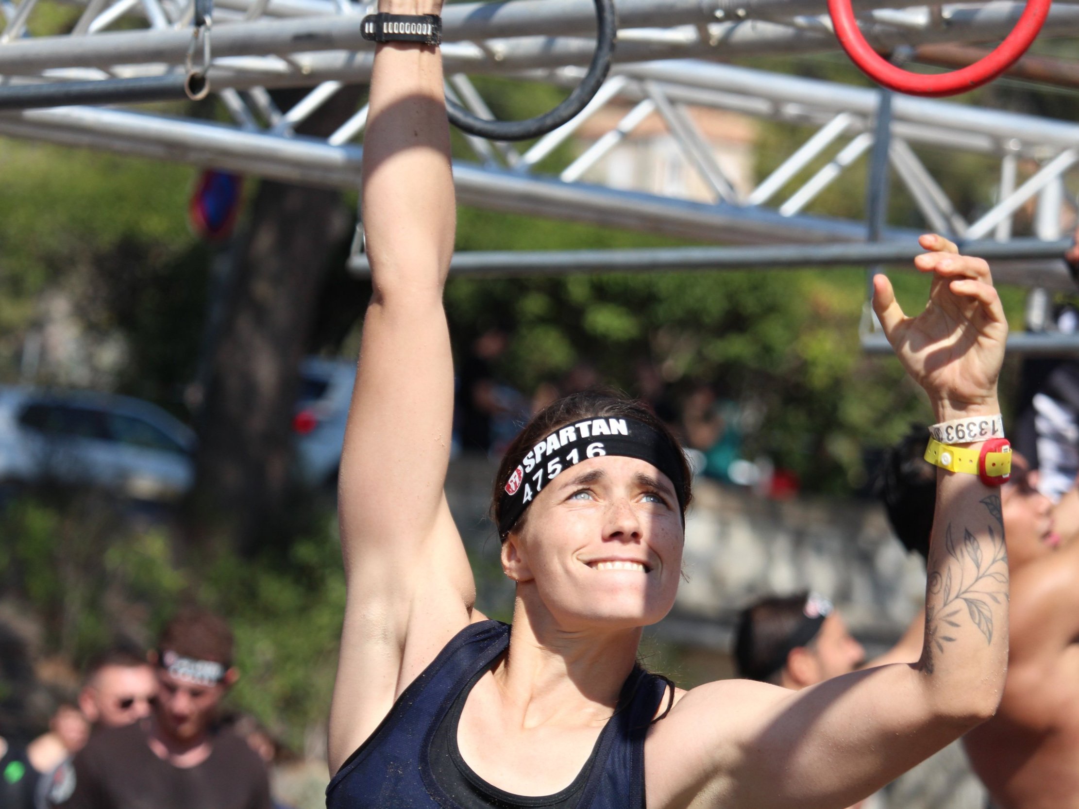 Une femme participe à une compétition de course d'obstacles, grimpe une structure métallique, portant un bandeau noir avec le mot 'SPARTAN', un bracelet jaune, et a un tatouage floral sur le bras.