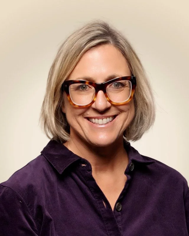 A woman smiling, wearing large black and brown glasses, a black sweater, and a black top, against a plain off-white background.