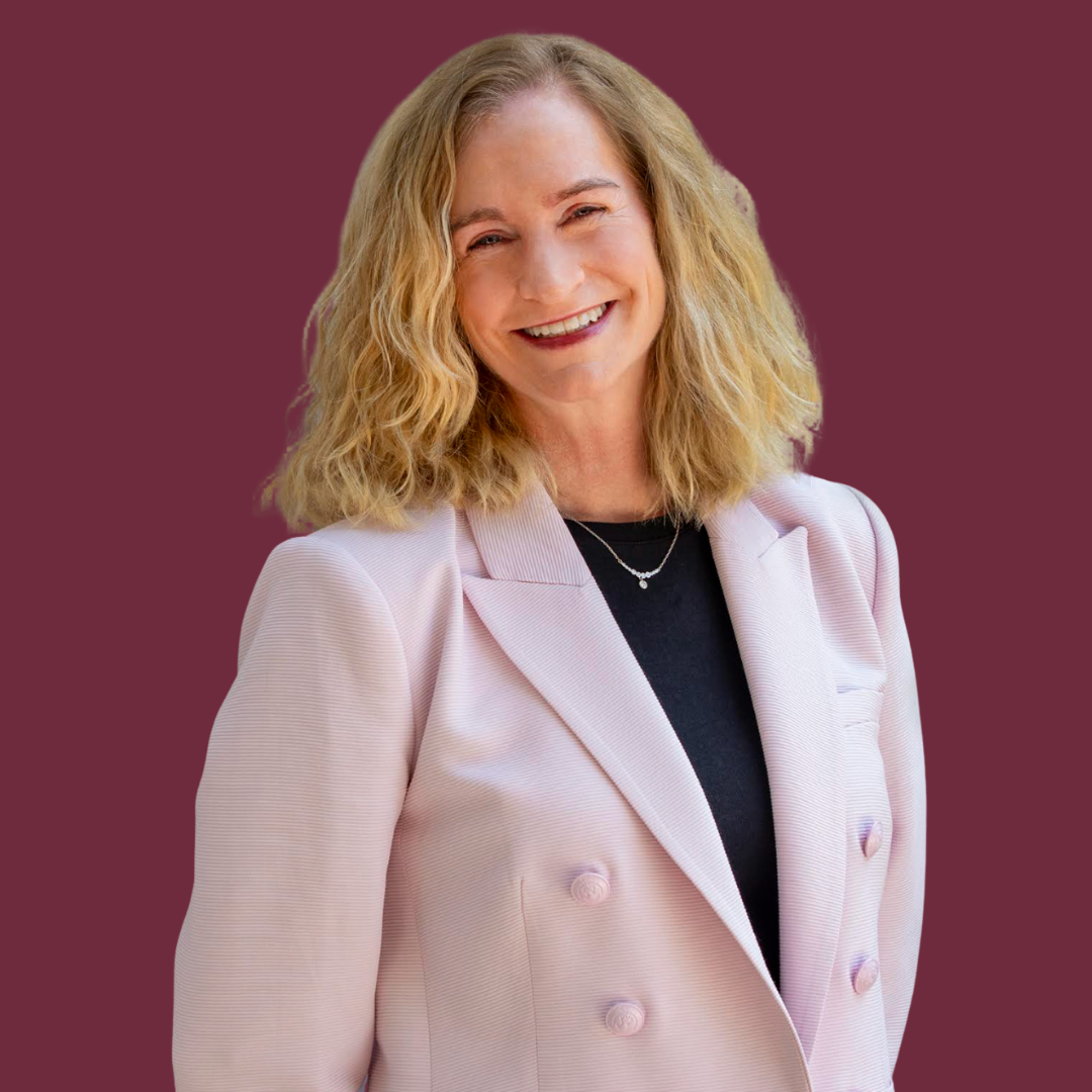 A woman with blonde, wavy hair smiling, wearing a light pink blazer over a black top, with a simple necklace, against a solid maroon background.