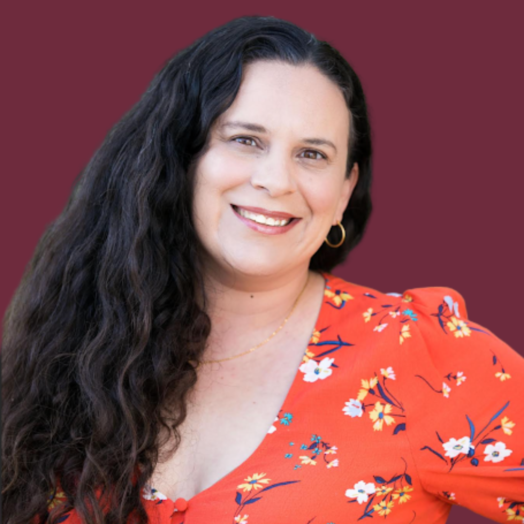 A woman with long, curly black hair, smiling, wearing a red floral top, with gold hoop earrings, a gold necklace, and a mauve background.
