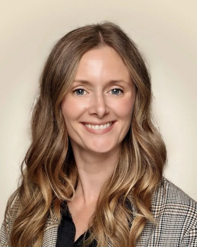 Headshot of a woman with long, wavy light brown hair, blue eyes, smiling, wearing a black top and a checked blazer against a plain background.