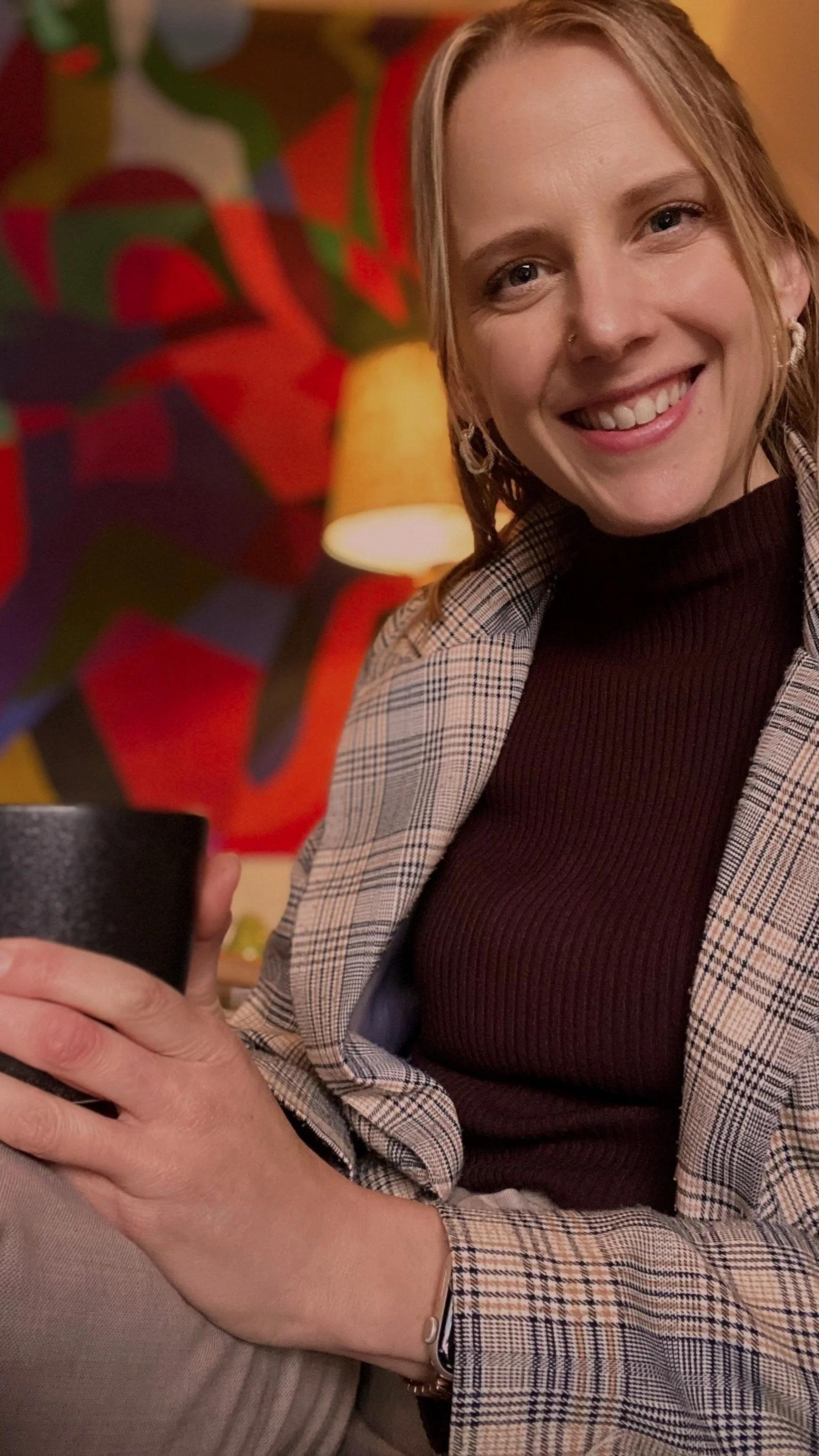 A woman with blonde hair smiling, holding a black cup, sitting indoors with a colorful, abstract painting and a yellow lamp in the background.