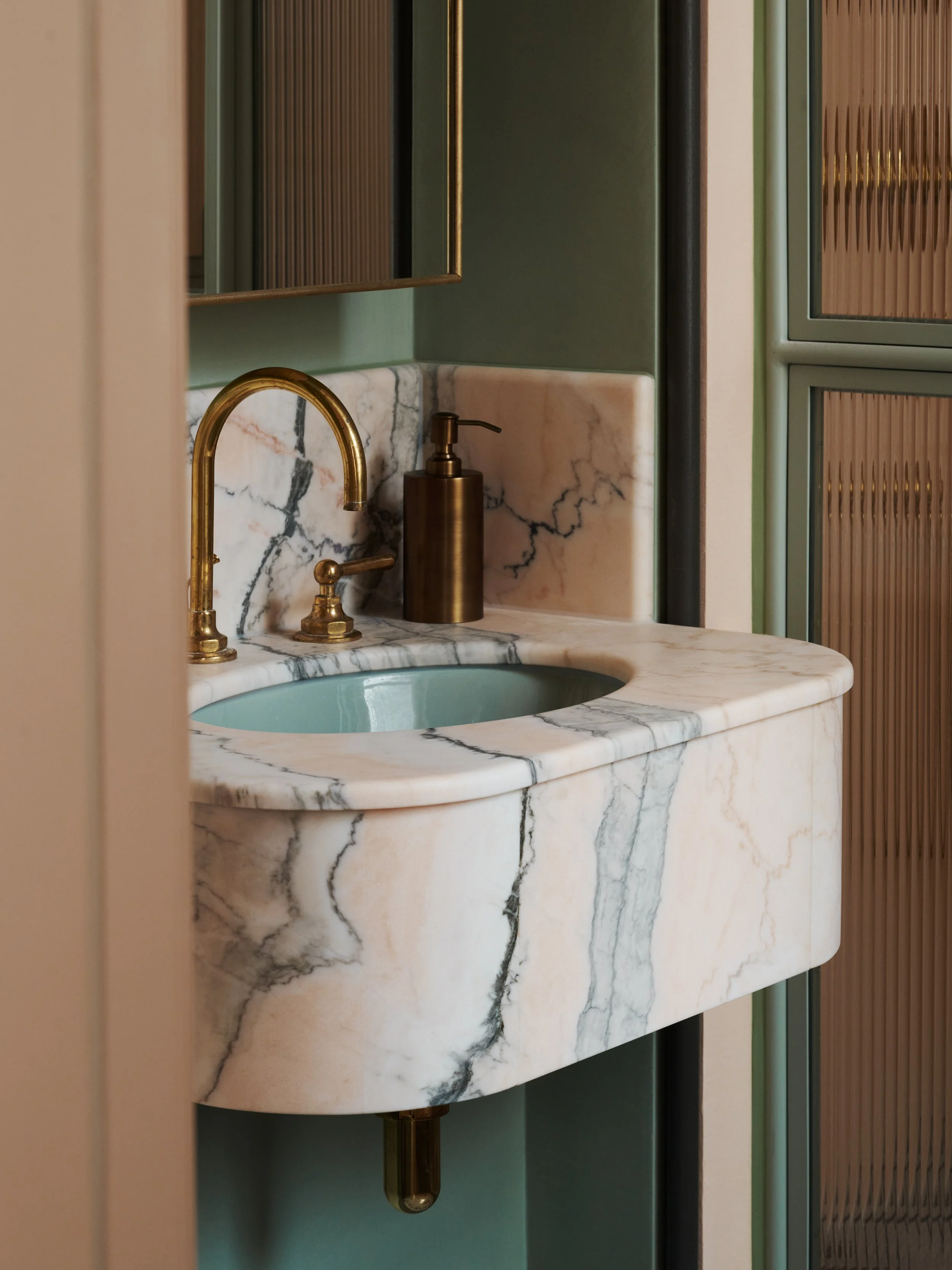 Close-up of a marble bathroom sink with a gold faucet, a brown soap dispenser, and a green wall with a mirror and textured cabinet.