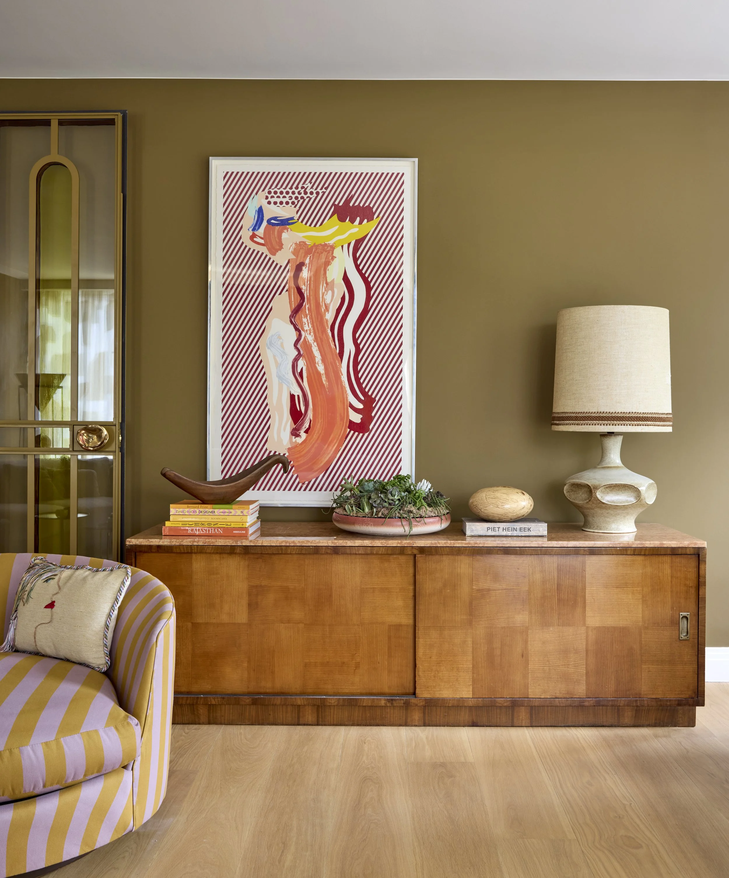 Interior of a living room featuring a wooden sideboard with decorative items, a large abstract art piece on a beige wall, a table lamp, a small plant, and a cushion on a striped armchair.