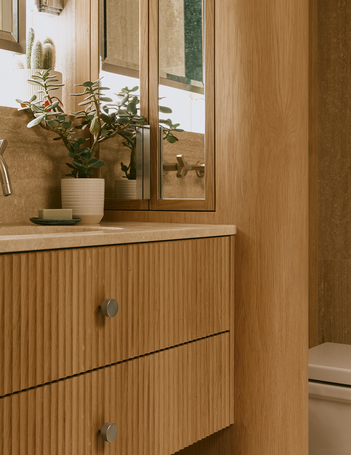 Close-up of a bathroom vanity with a wooden finish, a beige countertop, a potted plant, and a mirror reflecting the faucet.