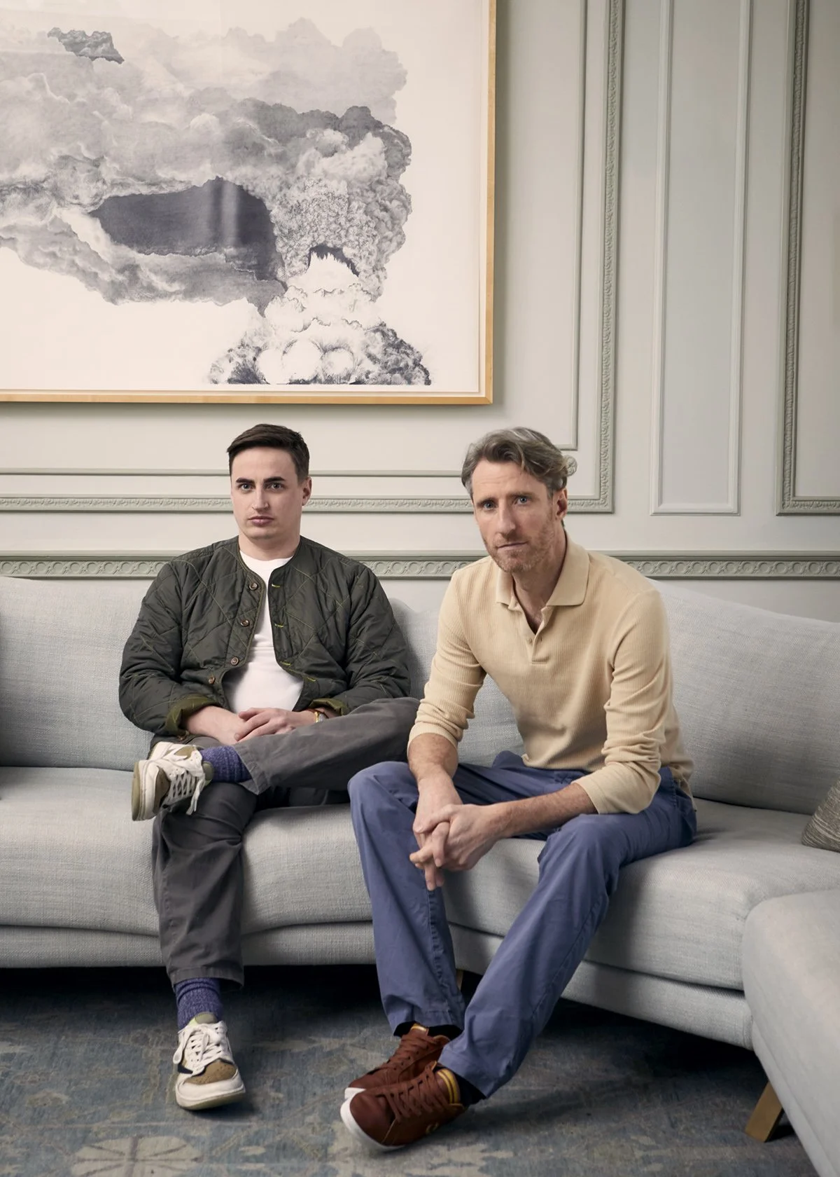 Two men sitting on a light-colored sofa in a room with wall paneling and a large wall art piece depicting clouds or smoke in a landscape style.