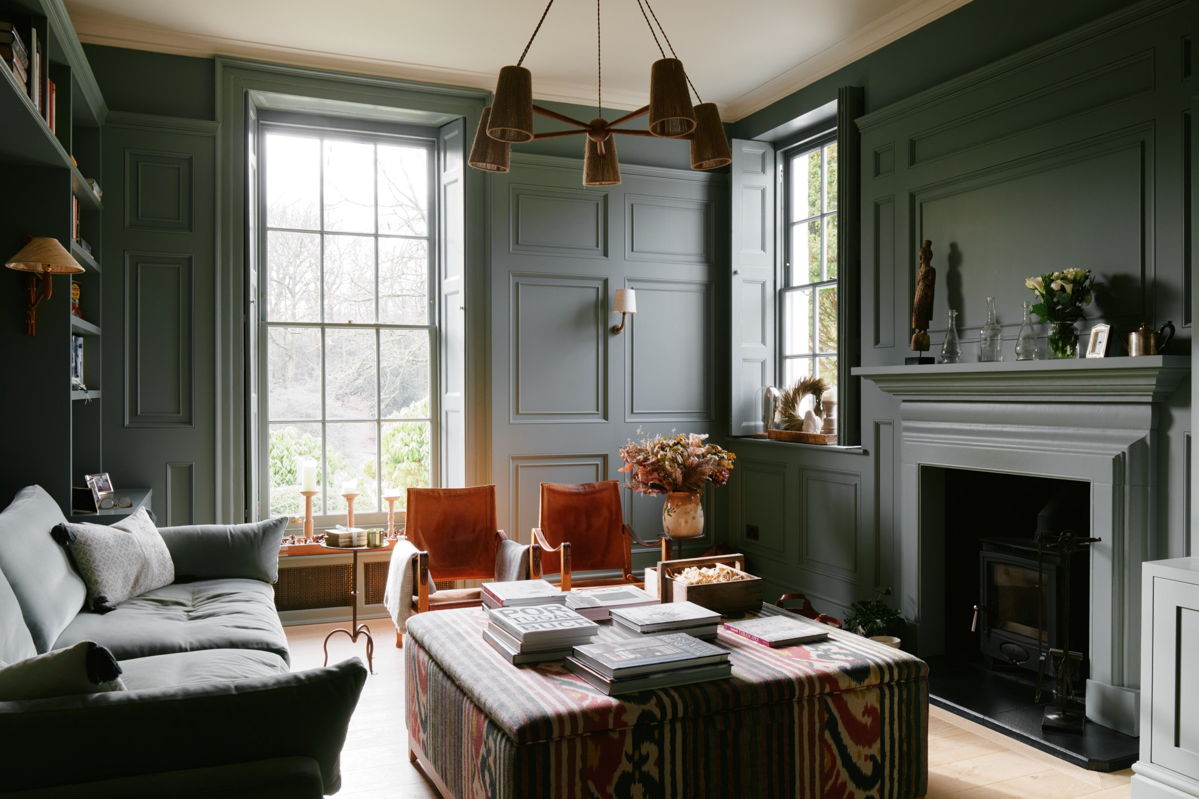 A cozy living room with green paneled walls, large windows, modern furniture, and a fireplace.