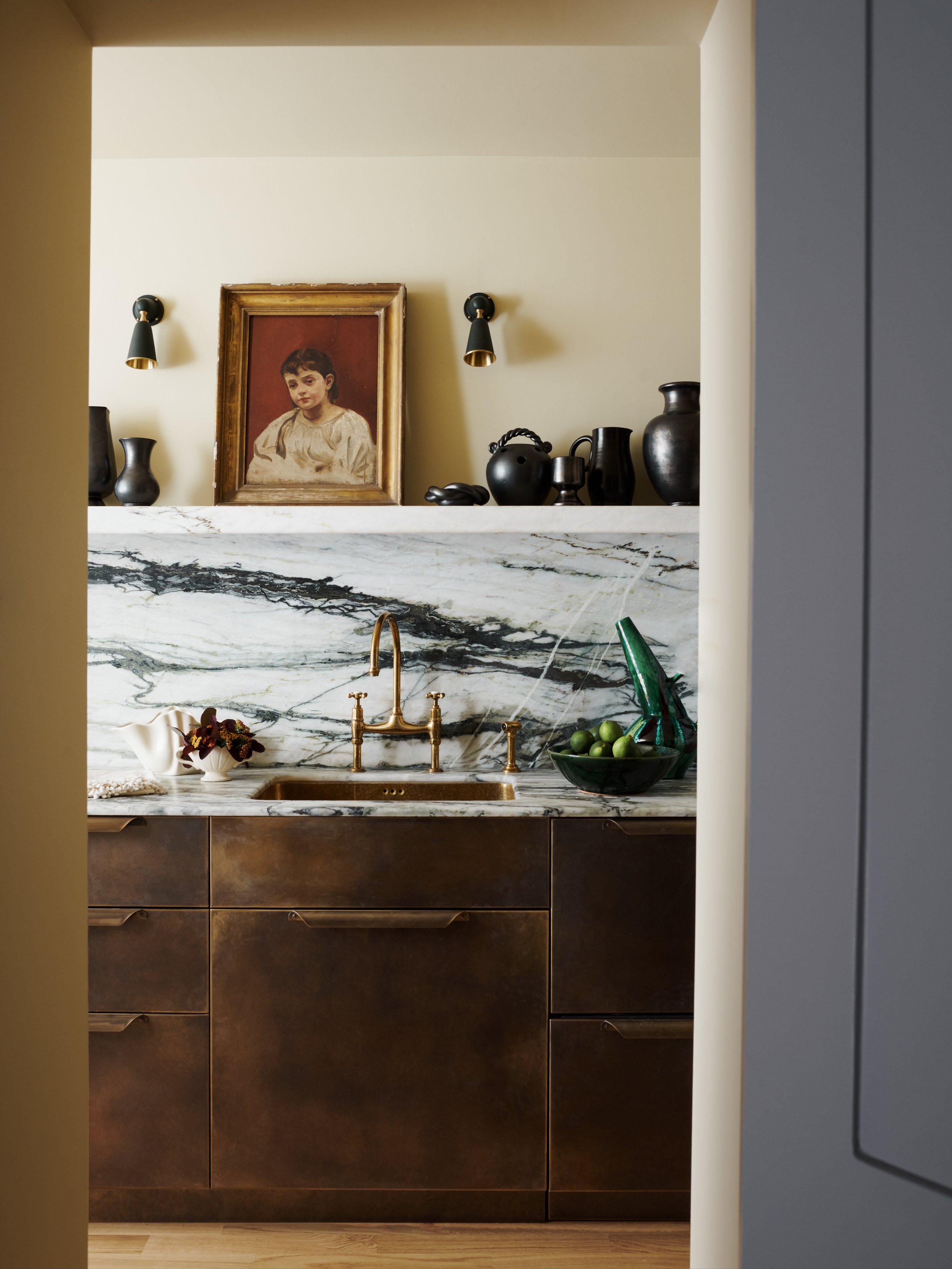 View of a kitchen counter with a marble backsplash, gold faucet, dark brown cabinets, a bowl of limes, and a framed portrait of a young girl on the wall behind. Black vases and small lights are also on the wall shelf above the counter.
