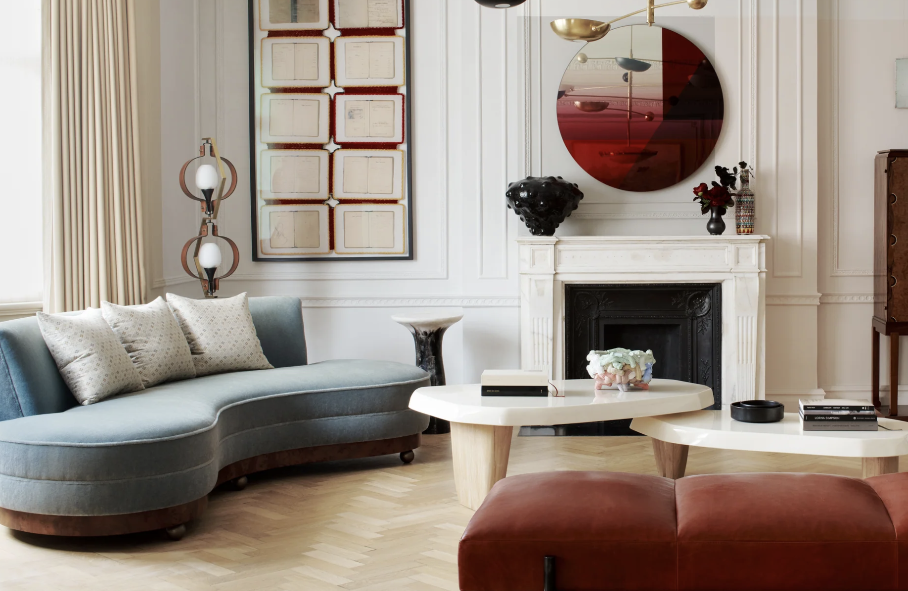 Living room with a light blue sofa with pillows, a white coffee table with decorative items, a white fireplace with black interior, a circular mirror with red design, framed artwork on the wall, a modern floor lamp, and various decorative objects.
