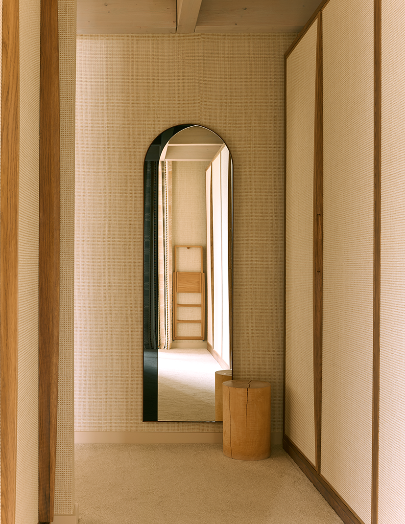 A room with a tall, narrow, arched mirror on a textured beige wall, with a wooden stool and a small wooden bench in front of the mirror, neutral-toned decor, and natural lighting.