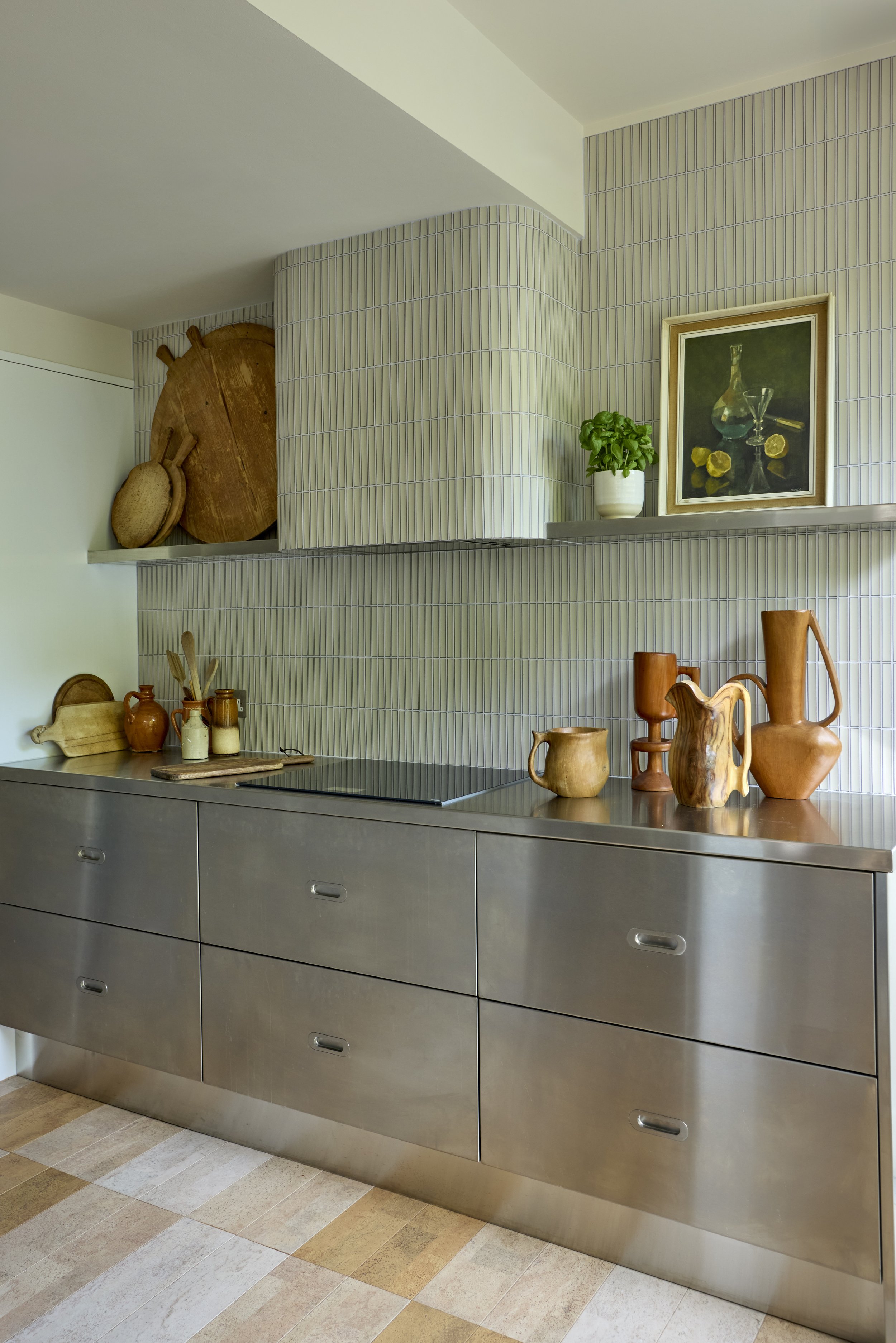 Modern kitchen with gray cabinetry, a black stovetop, wood cutting boards, pottery vases, and a framed painting on a light green tiled wall with a potted plant.