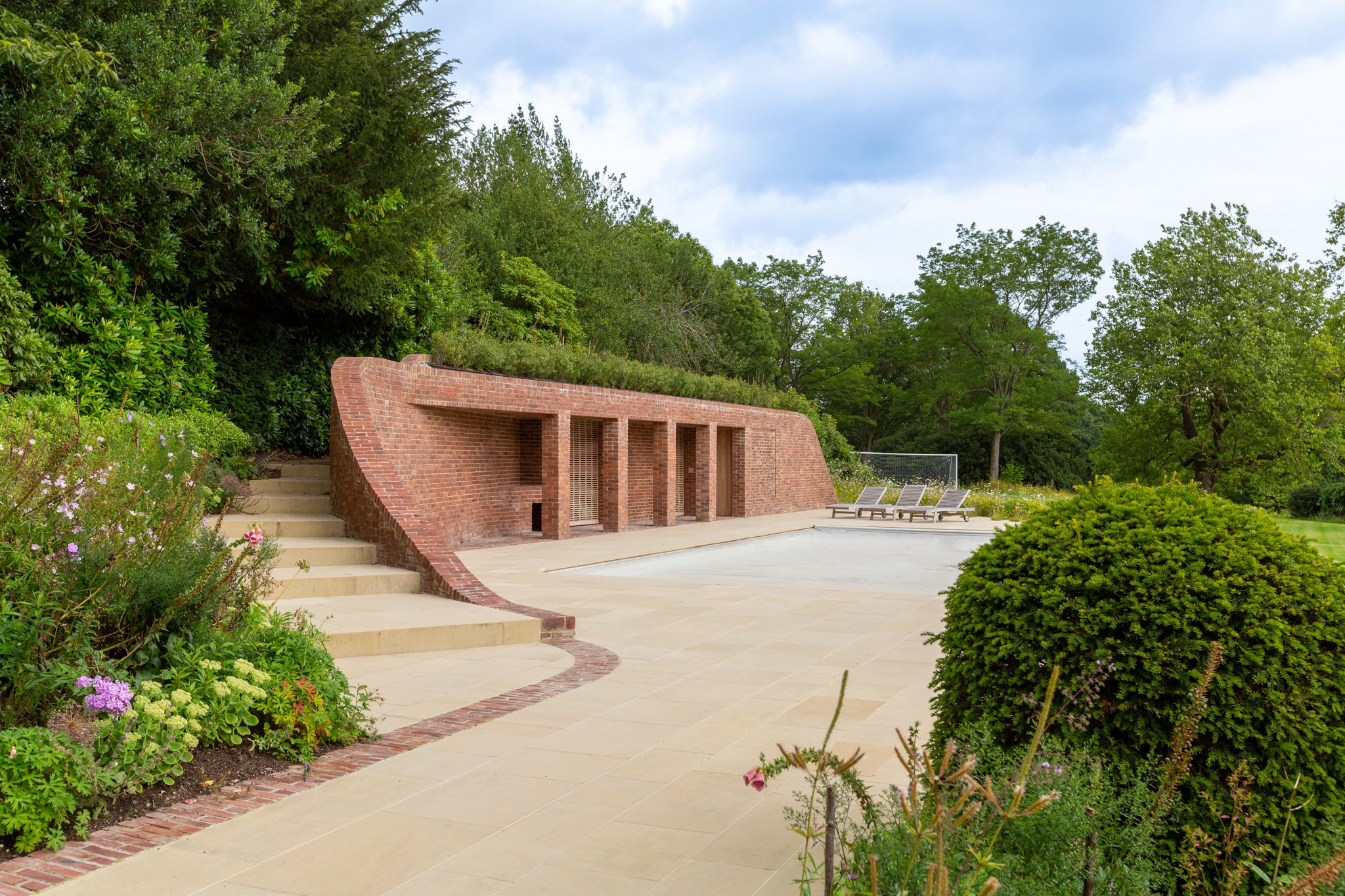 Empty swimming pool area with three lounge chairs on a paved patio, surrounded by greenery, a brick wall with archways, and steps leading up to a lush garden.