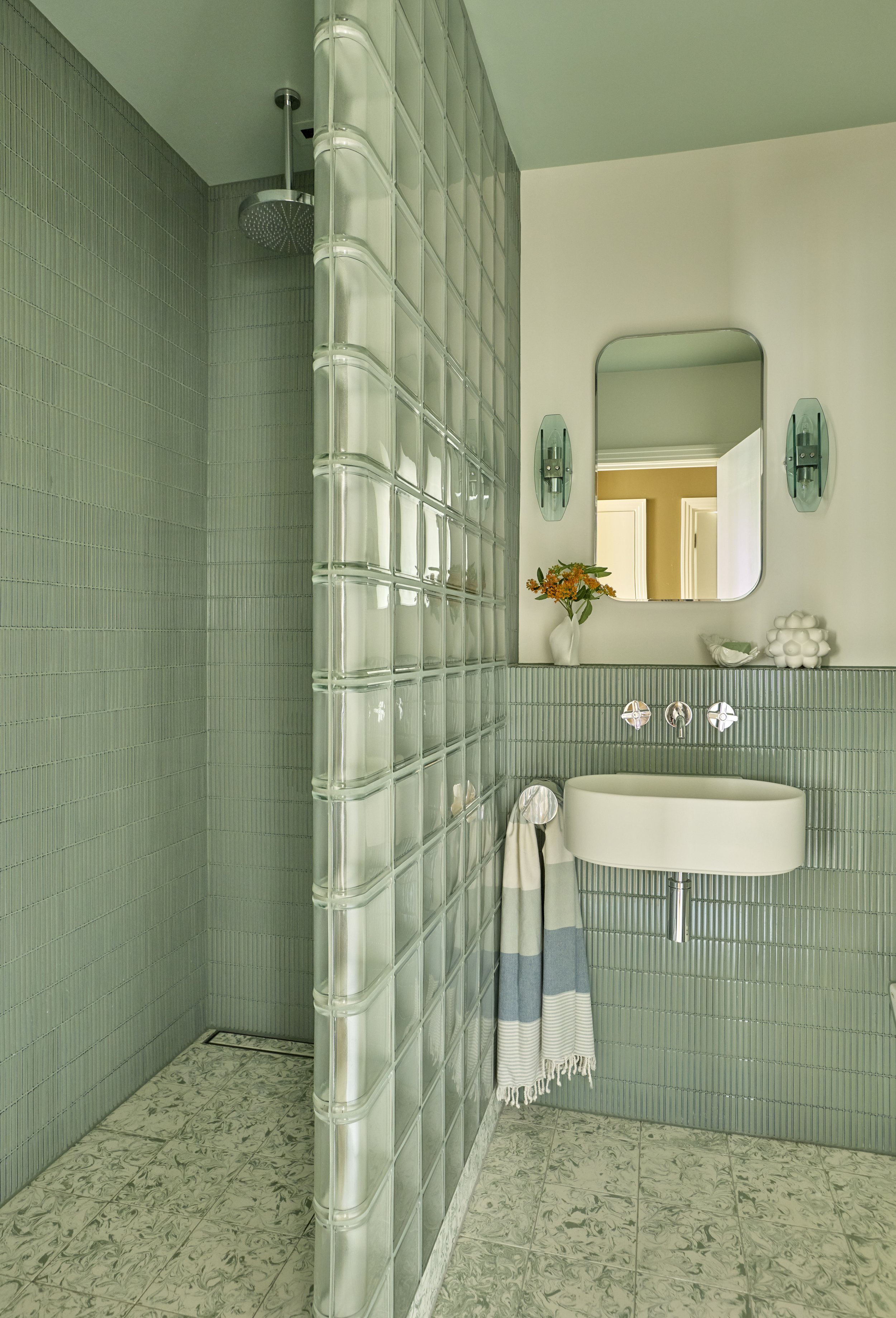 Bathroom with green tile walls, glass block shower wall, white sink, oval mirror, green glass wall sconces, and decorative items on the shelf