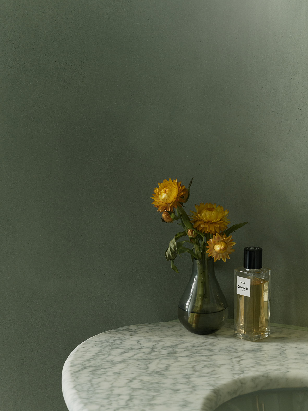 Yellow flowers in a clear glass vase and Chanel No. 22 perfume bottle on a white marble table against a green wall.