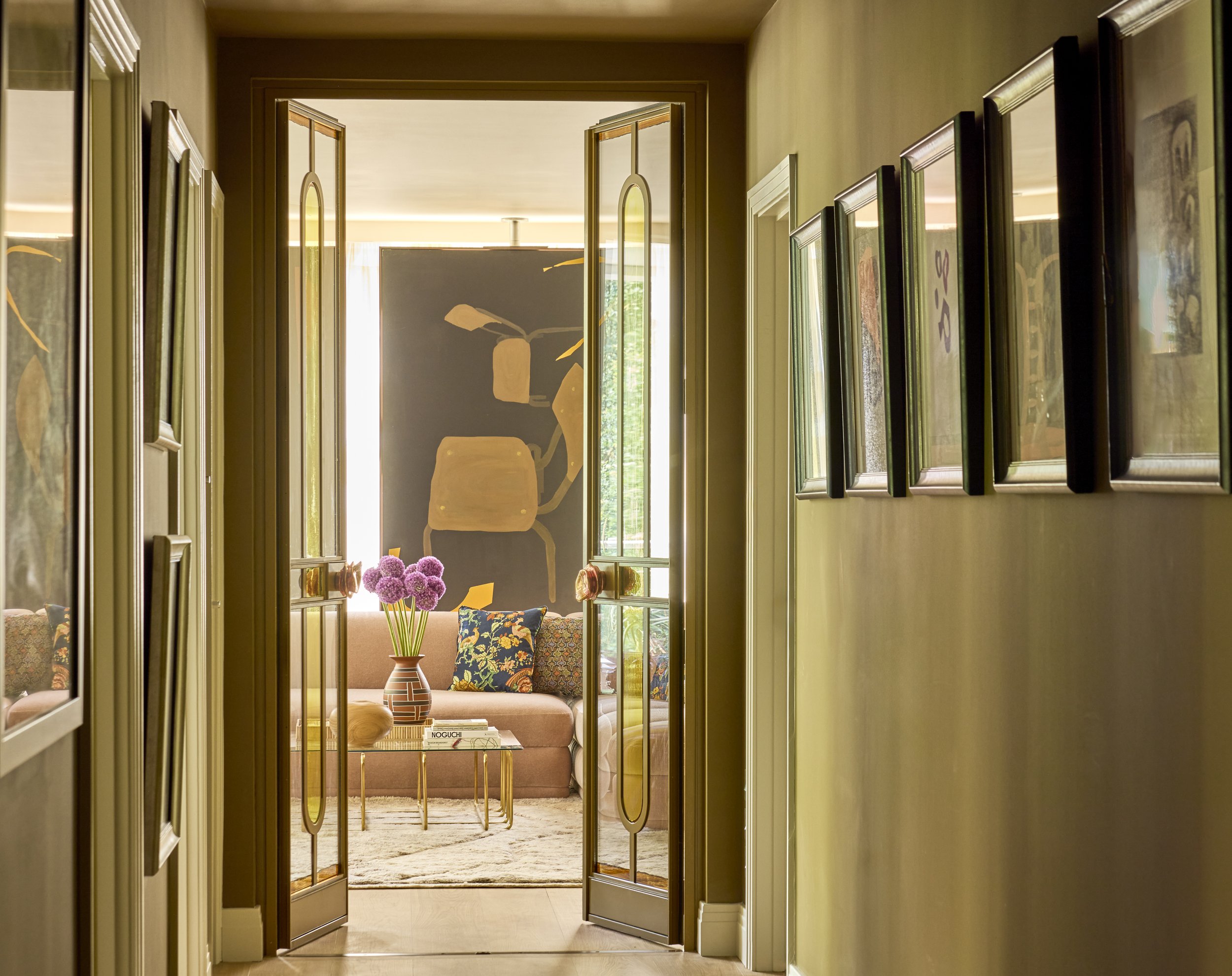 View through double glass doors into a sitting area with a pink sofa, colorful pillows, a vase with purple flowers on a glass table, and abstract artwork on the wall.