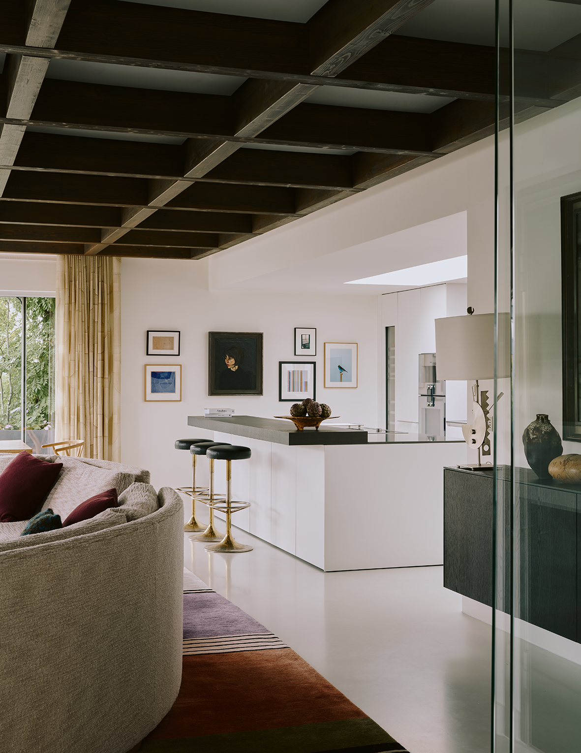 Modern living room with a curved sectional sofa, colorful pillows, a multicolored rug, a wall decorated with framed art, black barstools with gold bases, and a sleek white kitchen in the background.