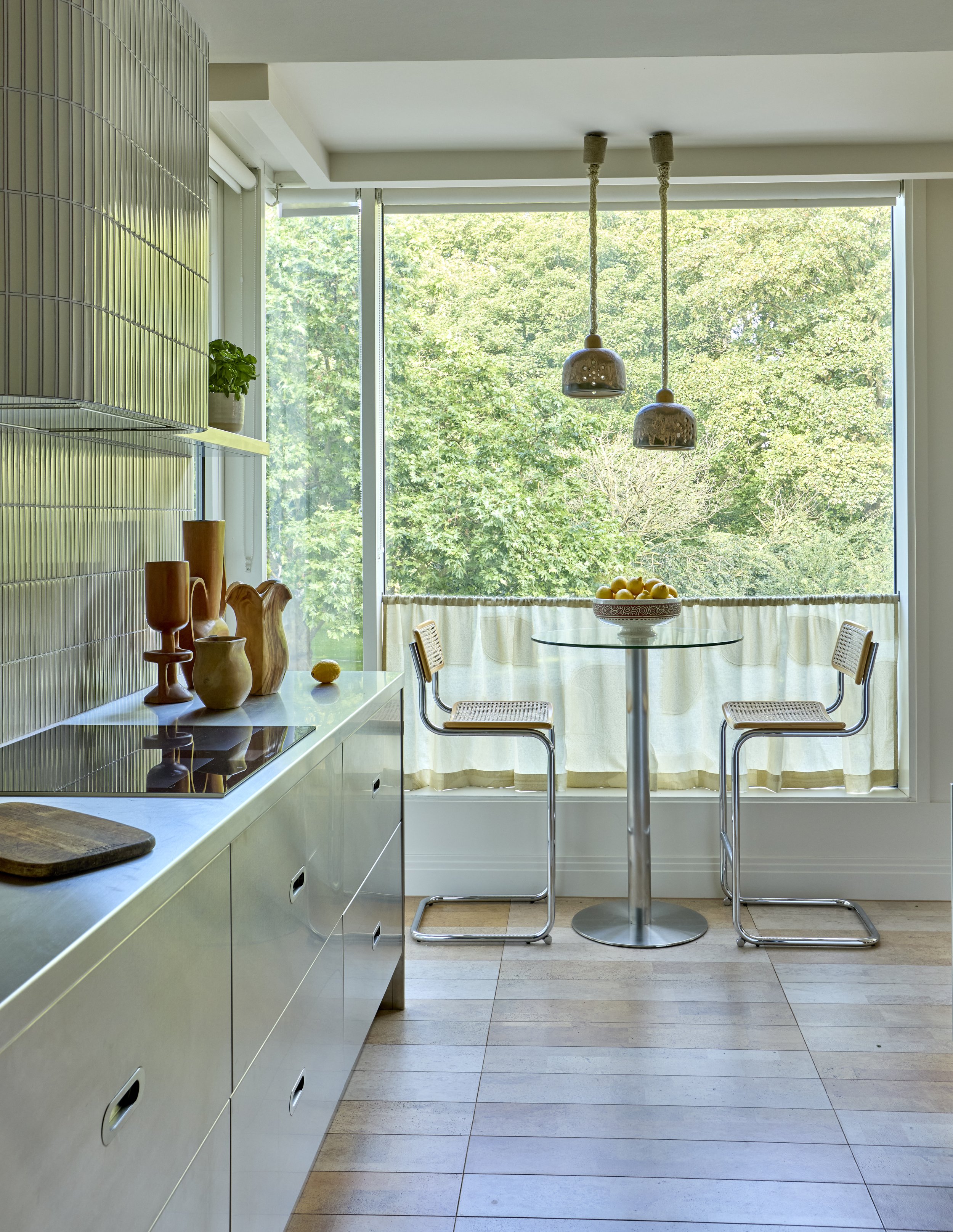 Modern kitchen with a large window, light-colored cabinets, decorative vases, and a small round table with two chairs, overlooking a lush green outdoor view.