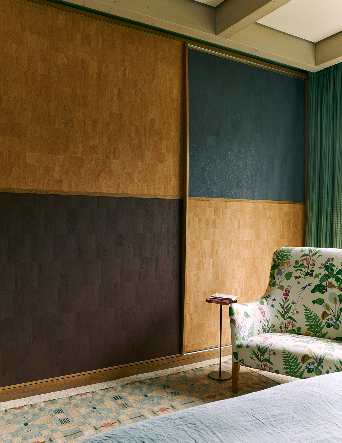 Interior of a room featuring a colorful floral-patterned armchair, a small side table with a book, a carpet with geometric design, green curtains, and a wall with multicolored wooden panels.