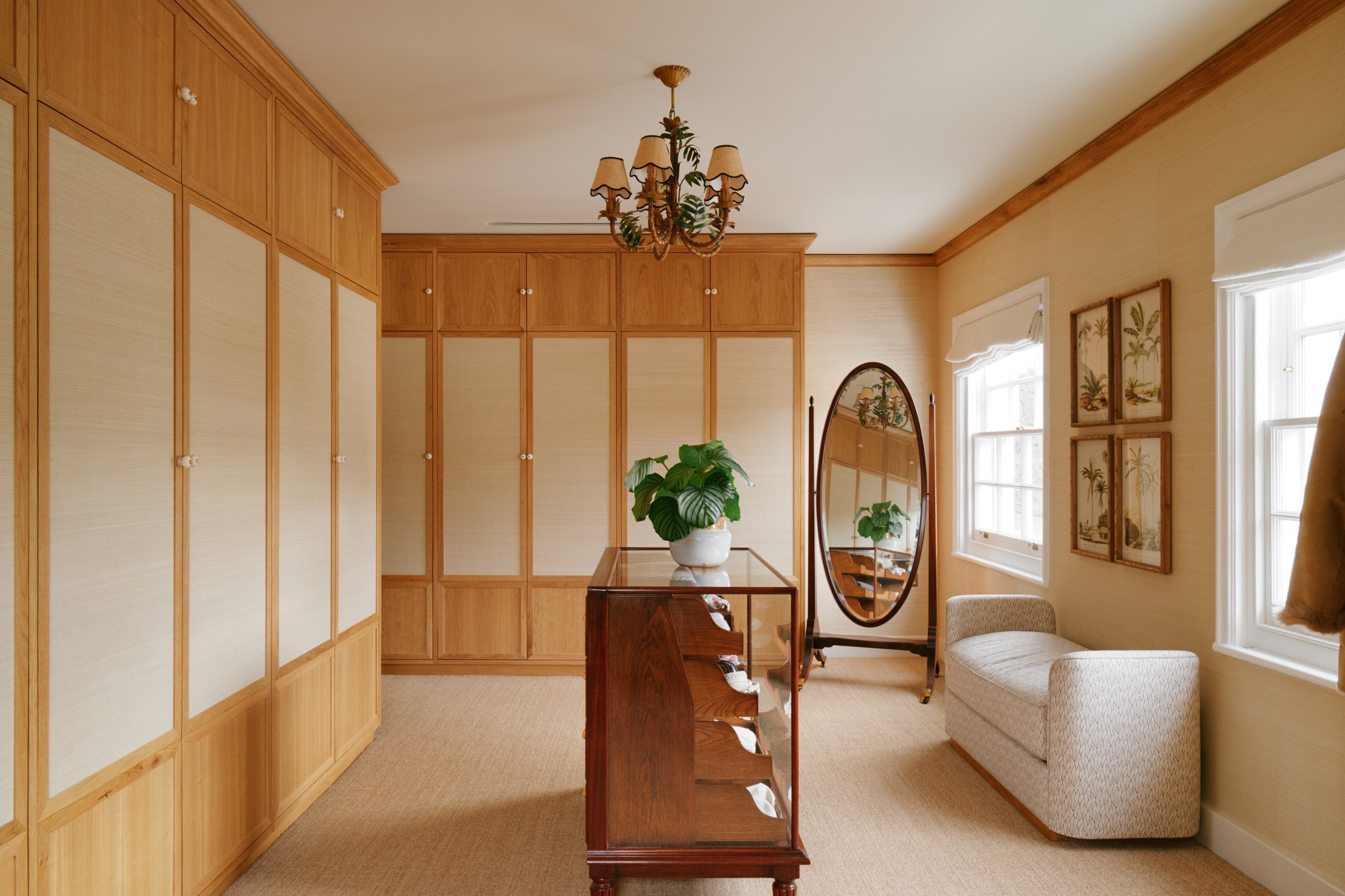 A cozy room with built-in wooden cabinets, a glass display case with decorative plates, a potted plant, a large wall mirror, a cushioned chaise lounge, and framed tropical art on the walls under natural light from windows.